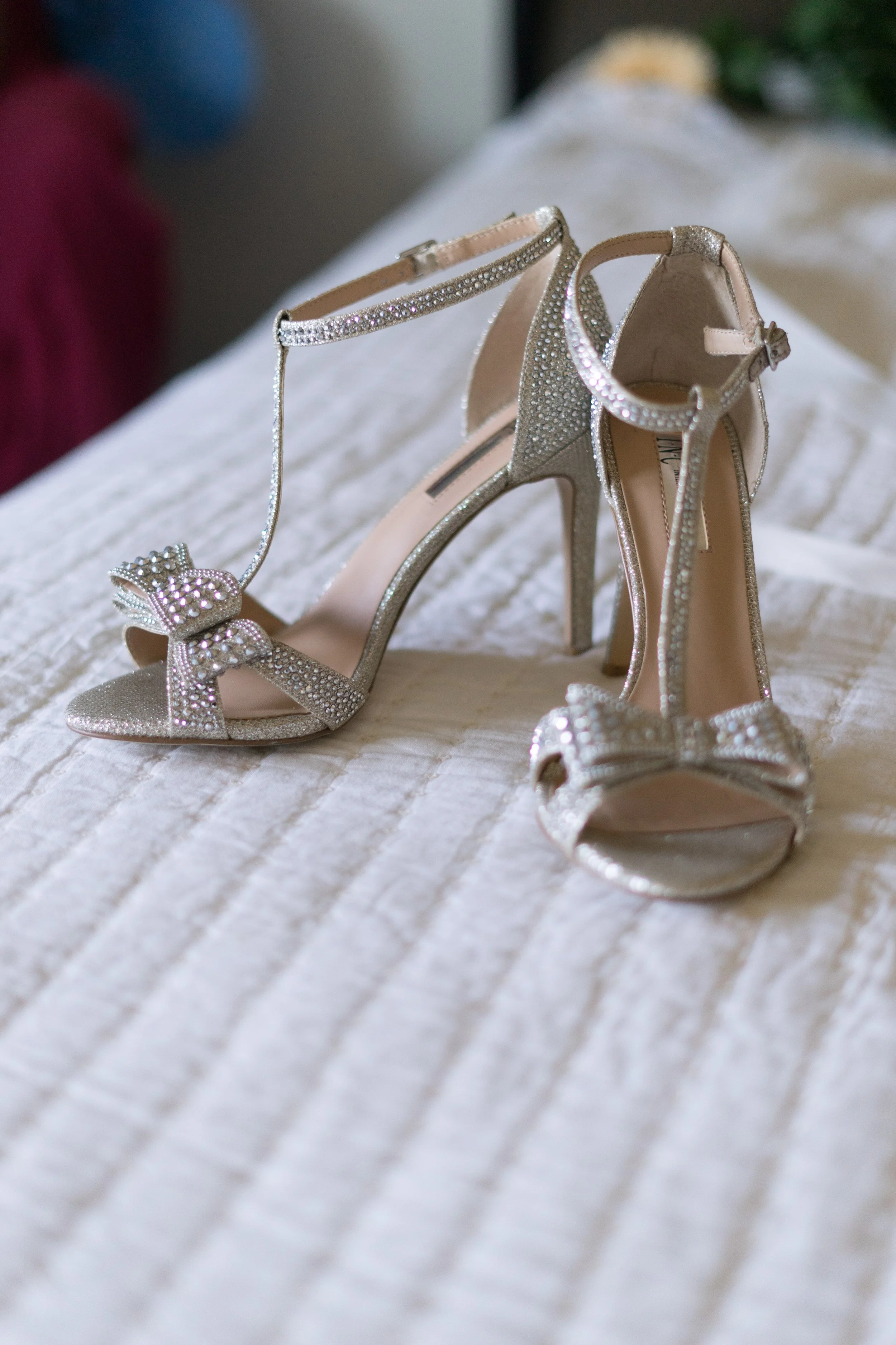 Silver high-heeled sandals decorated with rhinestones and a bow, placed on a white textured surface.