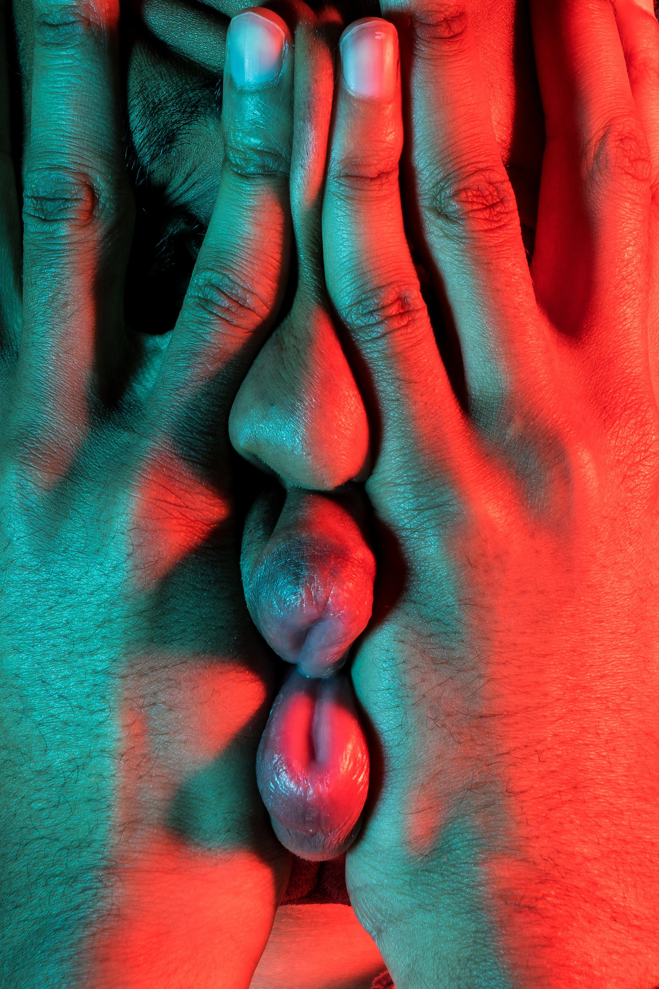 Close-up of two hands with manicured nails covering and pressing against a person's face with visible nose, lips, and chin, illuminated with red and teal lighting.