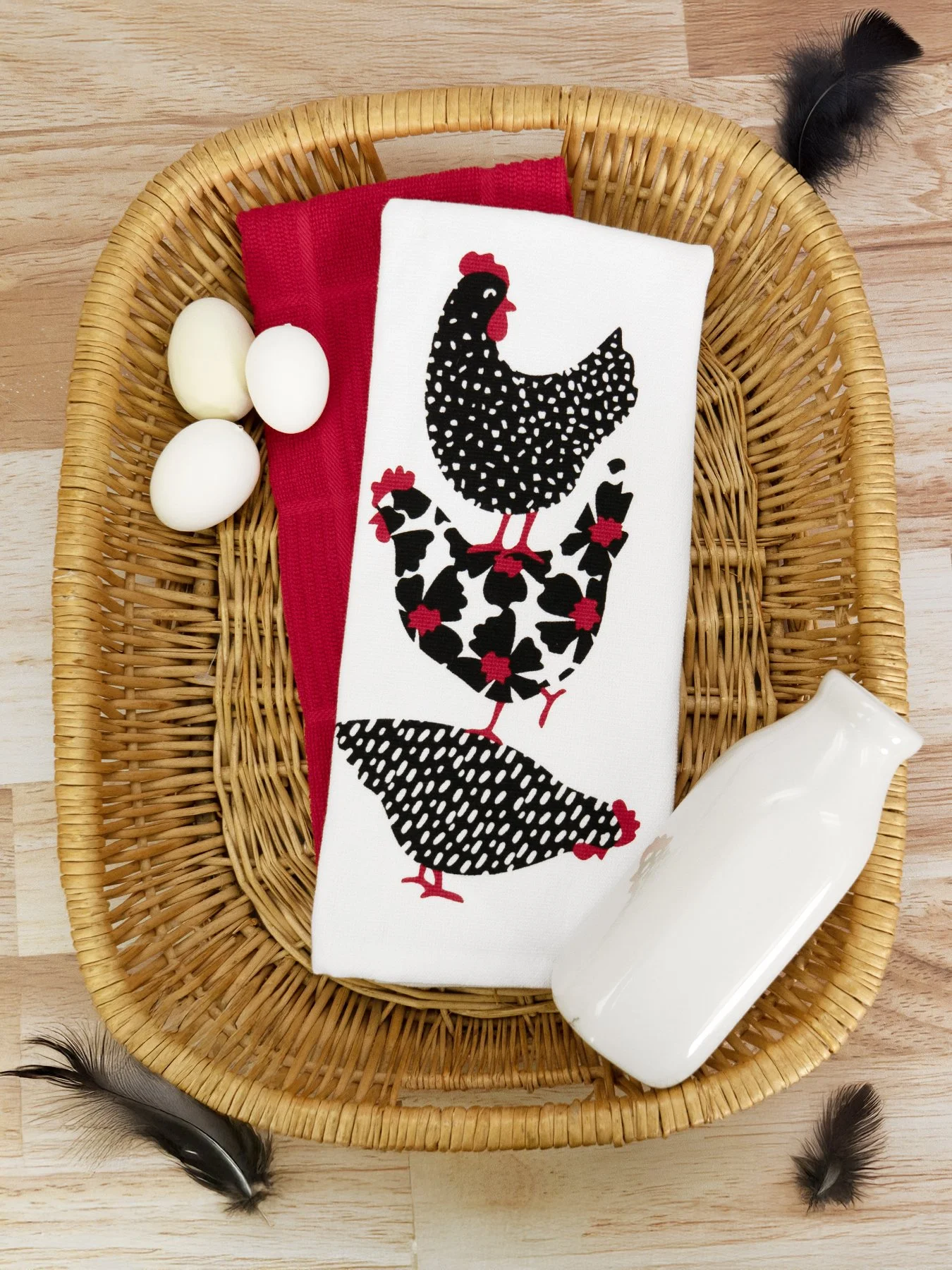 A wicker basket containing eggs, red cloth, a white cloth with a black rooster pattern, and a white ceramic dish, with black feathers around the basket on a wooden surface.