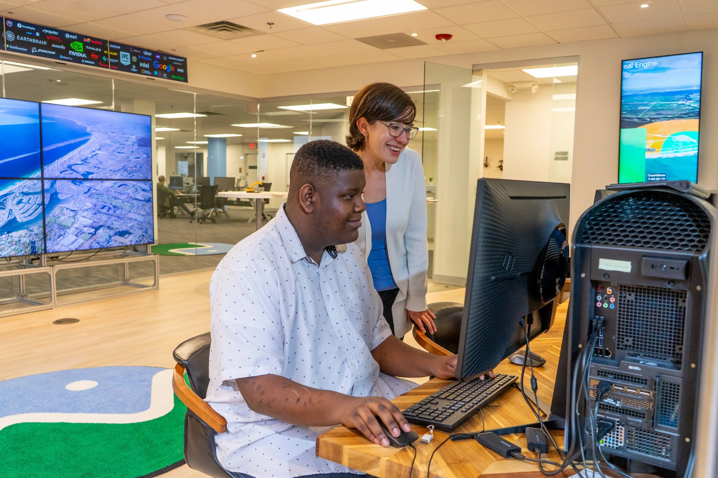 Two women in an office working at a desktop computer, with large screens displaying maps and data behind them.
