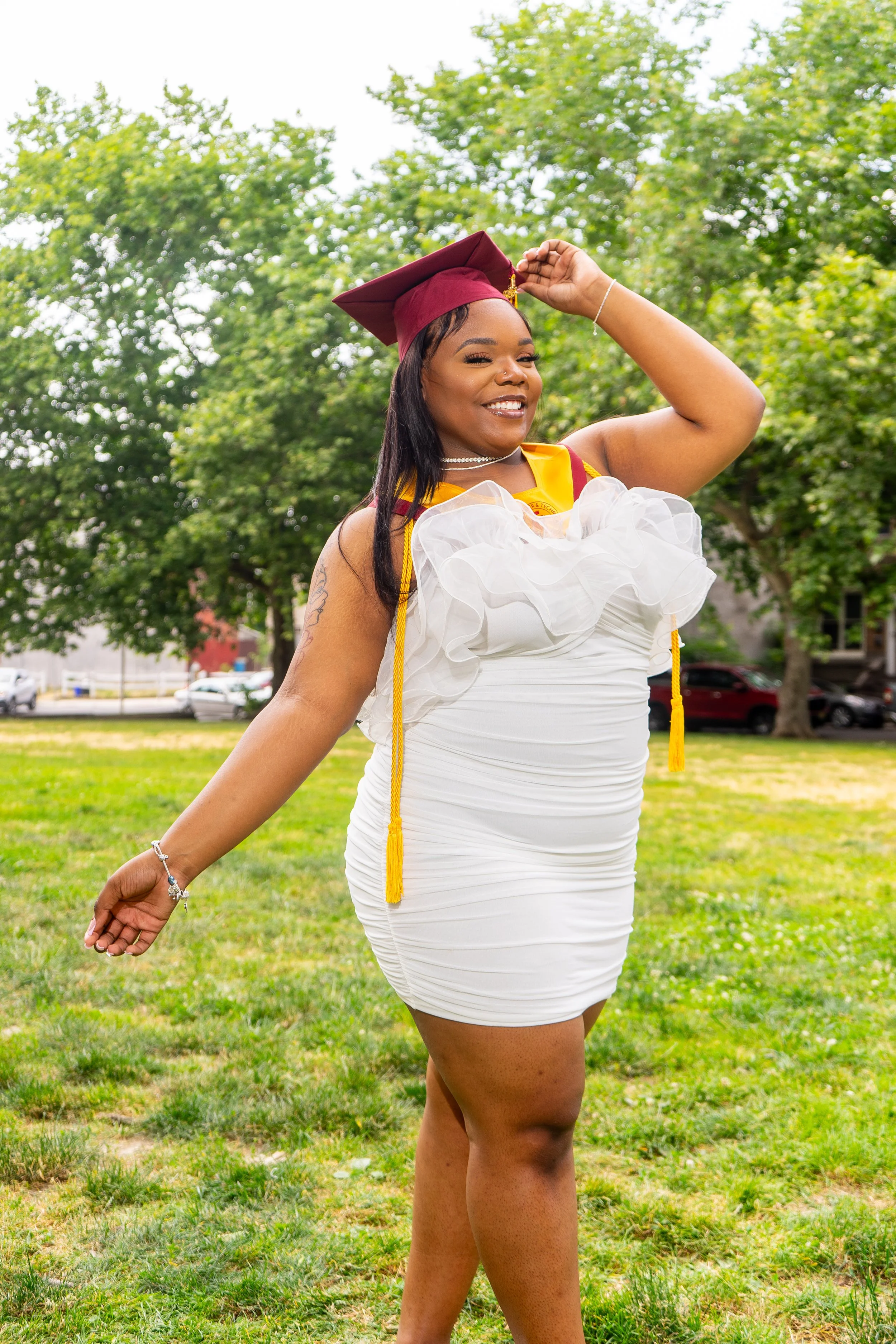 A young woman wearing a white dress and maroon graduation cap with yellow tassels, standing outdoors on a grassy area with trees and parked cars in the background. She is smiling, posing with her hand on her head.