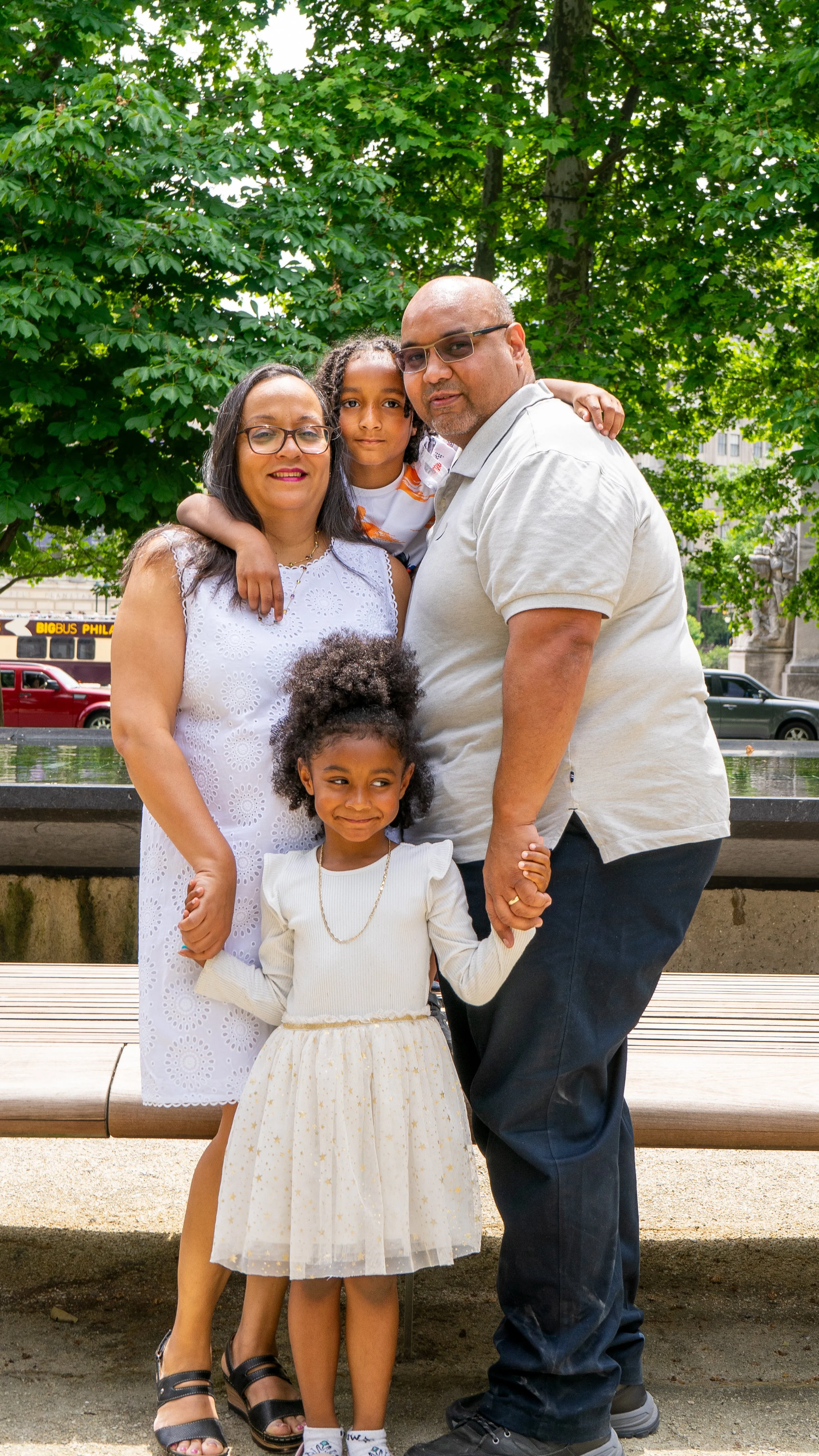 A family of five posing outdoors in front of green trees. The mother and father are holding hands, with two daughters standing in front and one son on the father's shoulders.