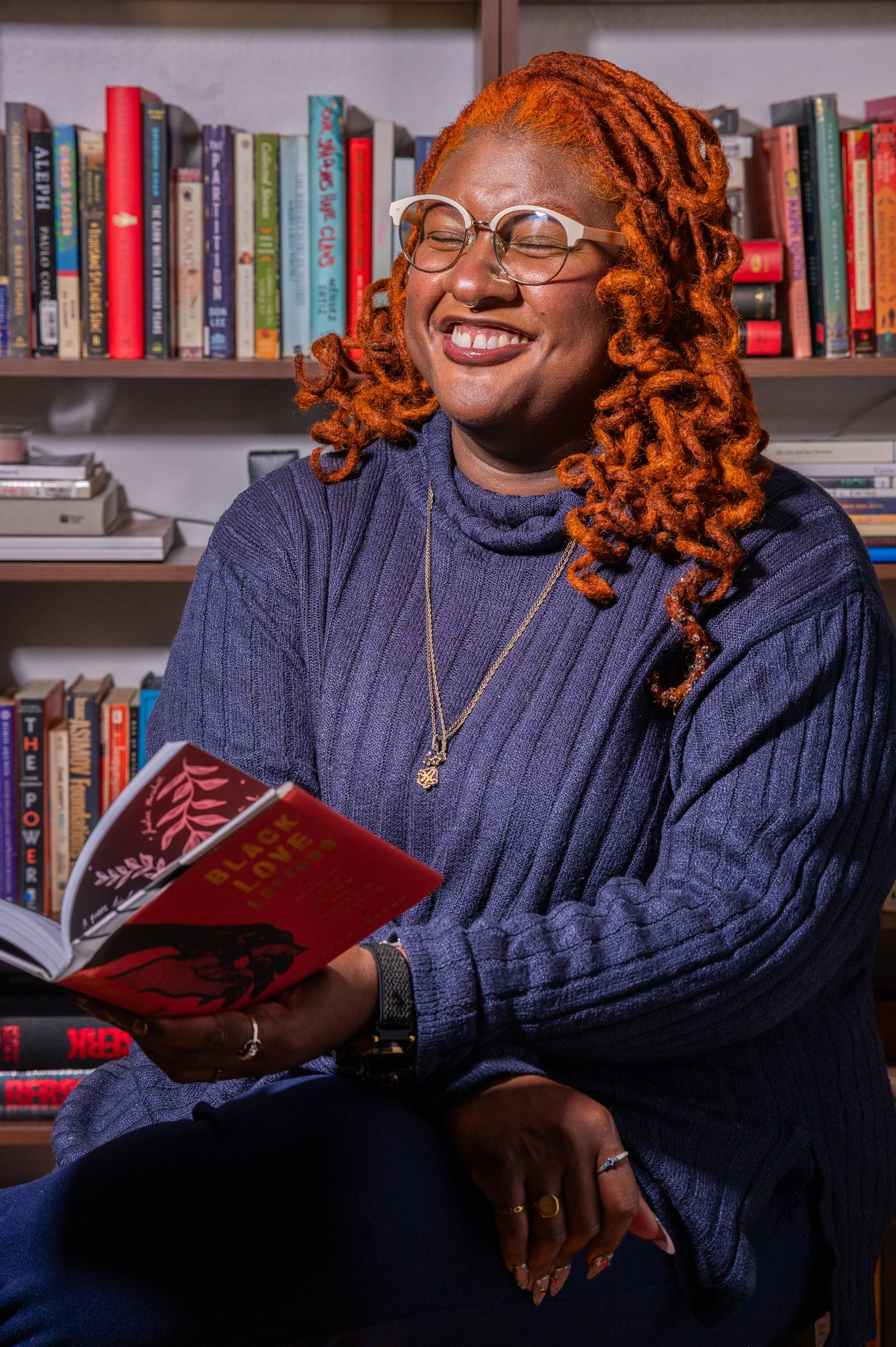 A woman with orange dreadlocks, glasses, and a dark purple sweater sitting in a bookstore, smiling with her eyes closed while holding a book titled 'Black Love'.