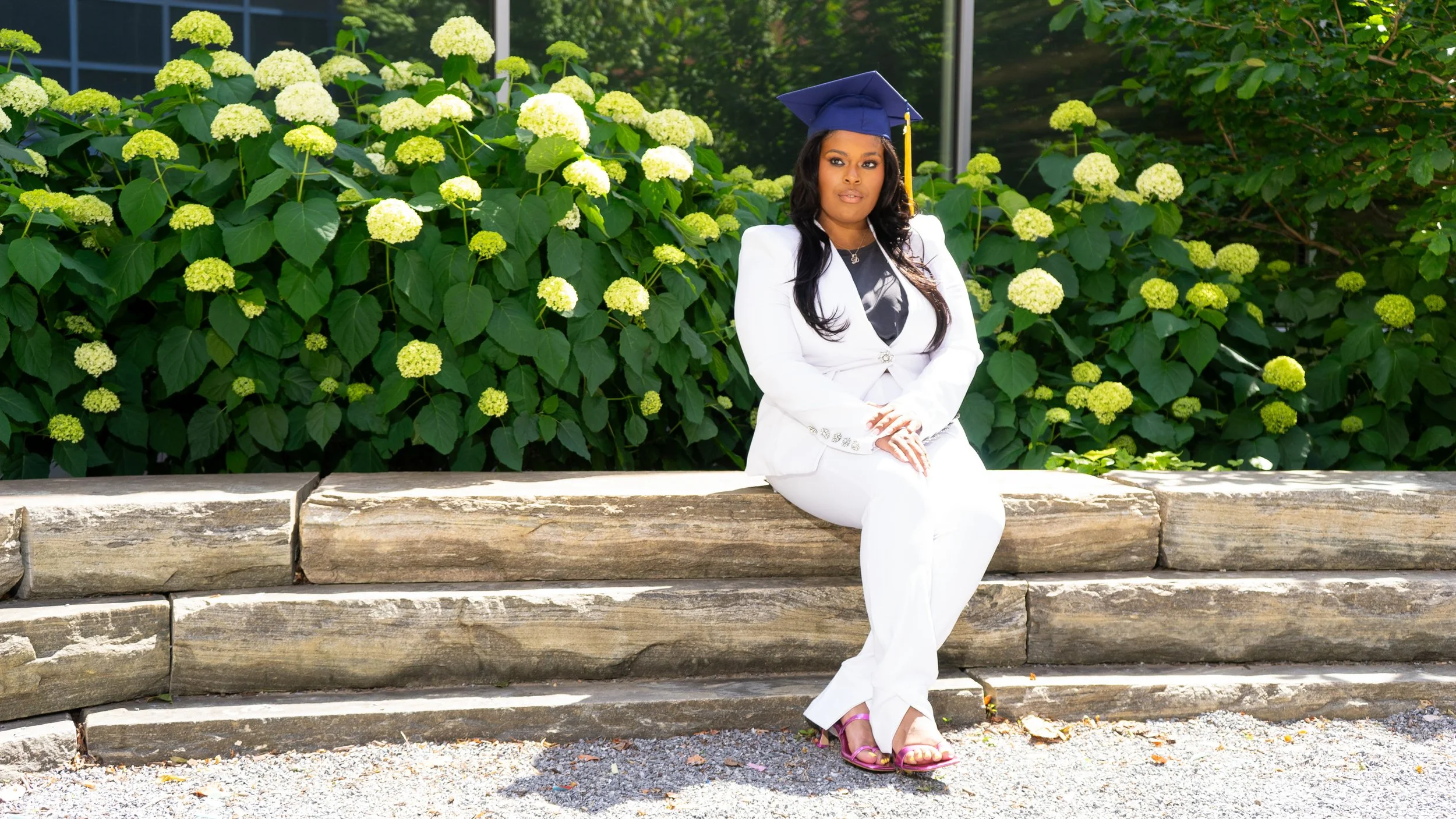 Woman wearing a white suit and graduation cap sitting on a stone ledge outdoors, with large green leafy plants and white hydrangea flowers in the background.