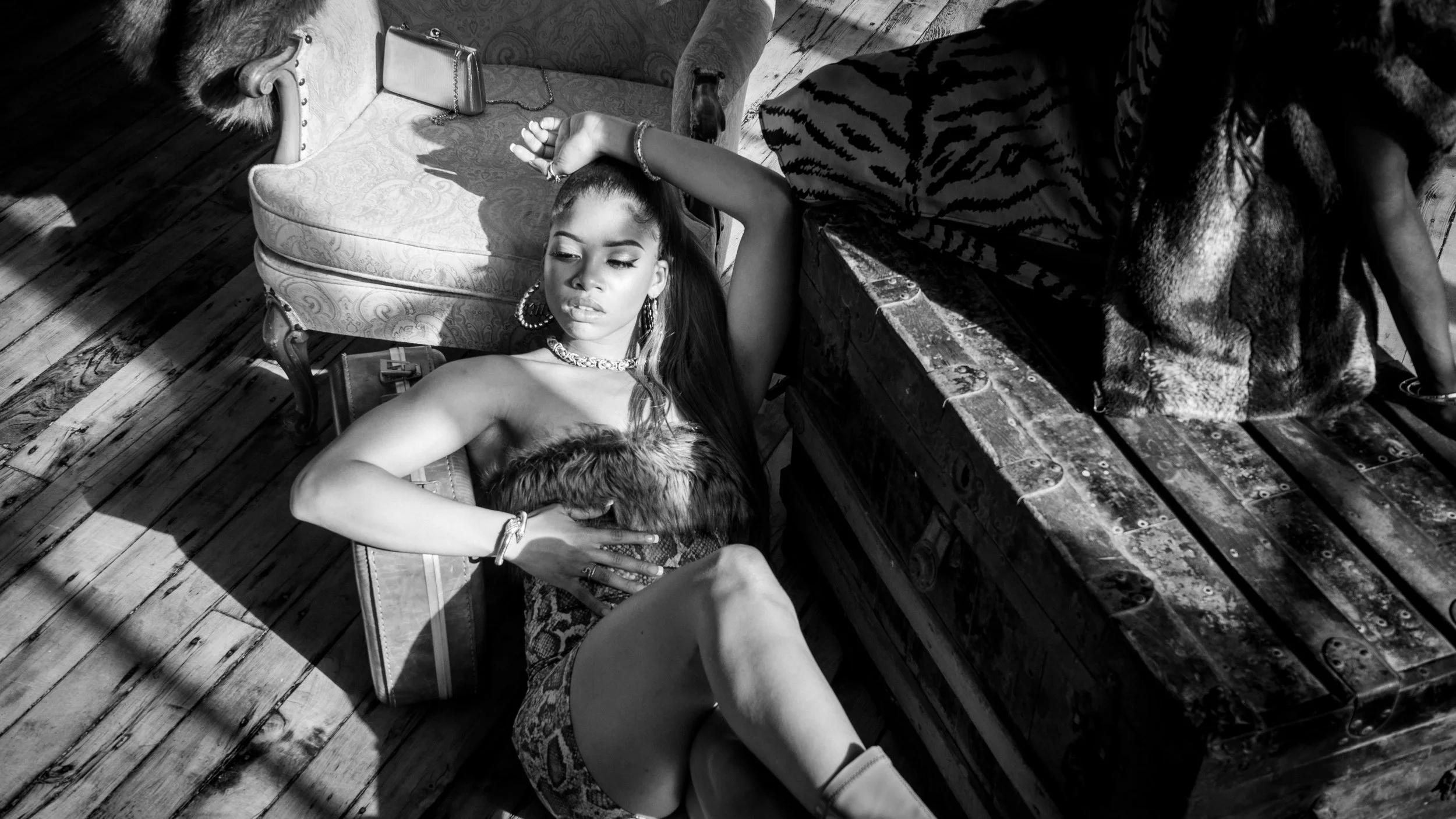 A woman with long hair, wearing large hoop earrings and jewelry, lays on the floor with her eyes closed, surrounded by vintage furniture, wooden floorboards, and a person partially visible on a nearby wooden surface.