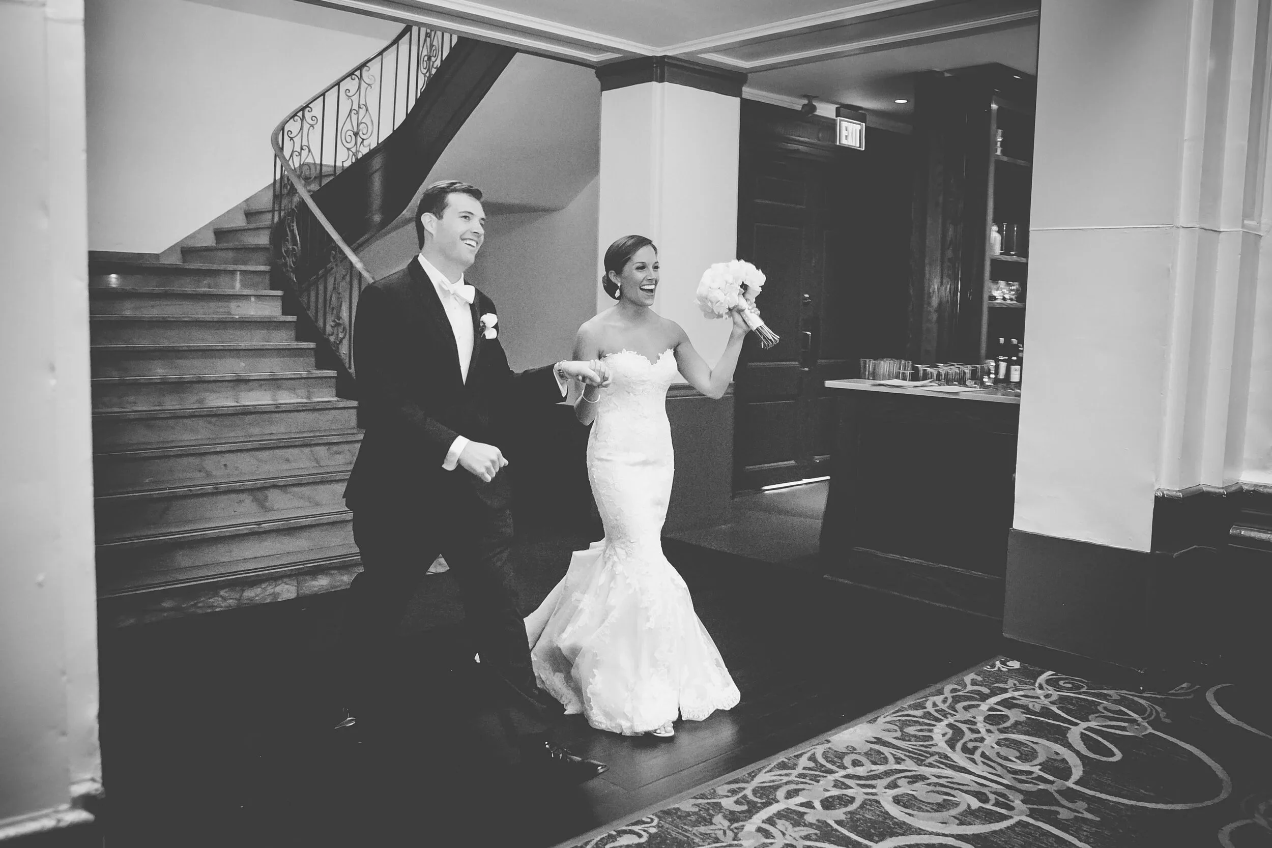 Black and white photo of a newlywed couple walking into a venue, smiling, with the bride holding a bouquet of flowers.