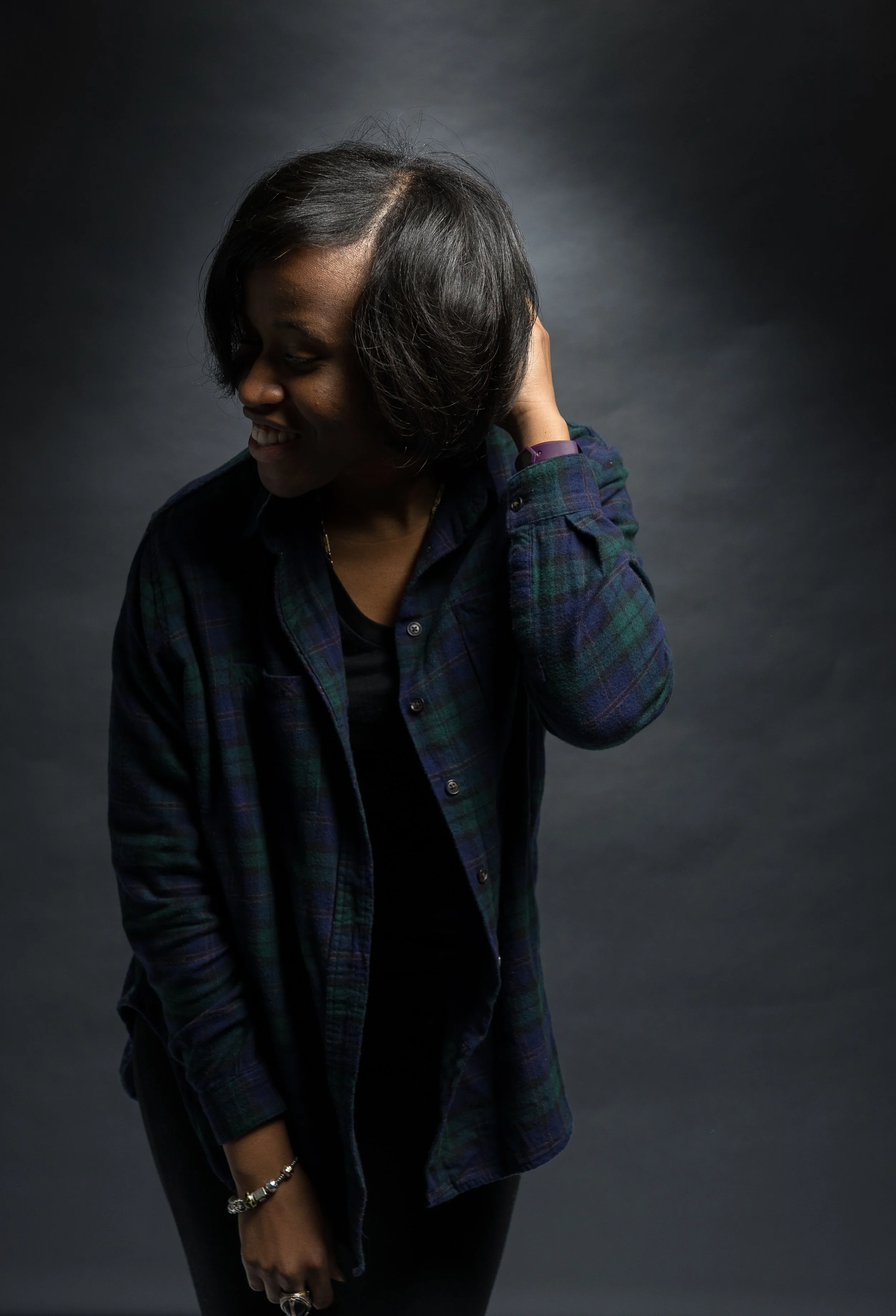 A woman with short black hair smiling and looking downward, wearing a dark plaid shirt and a black top, standing against a dark background.