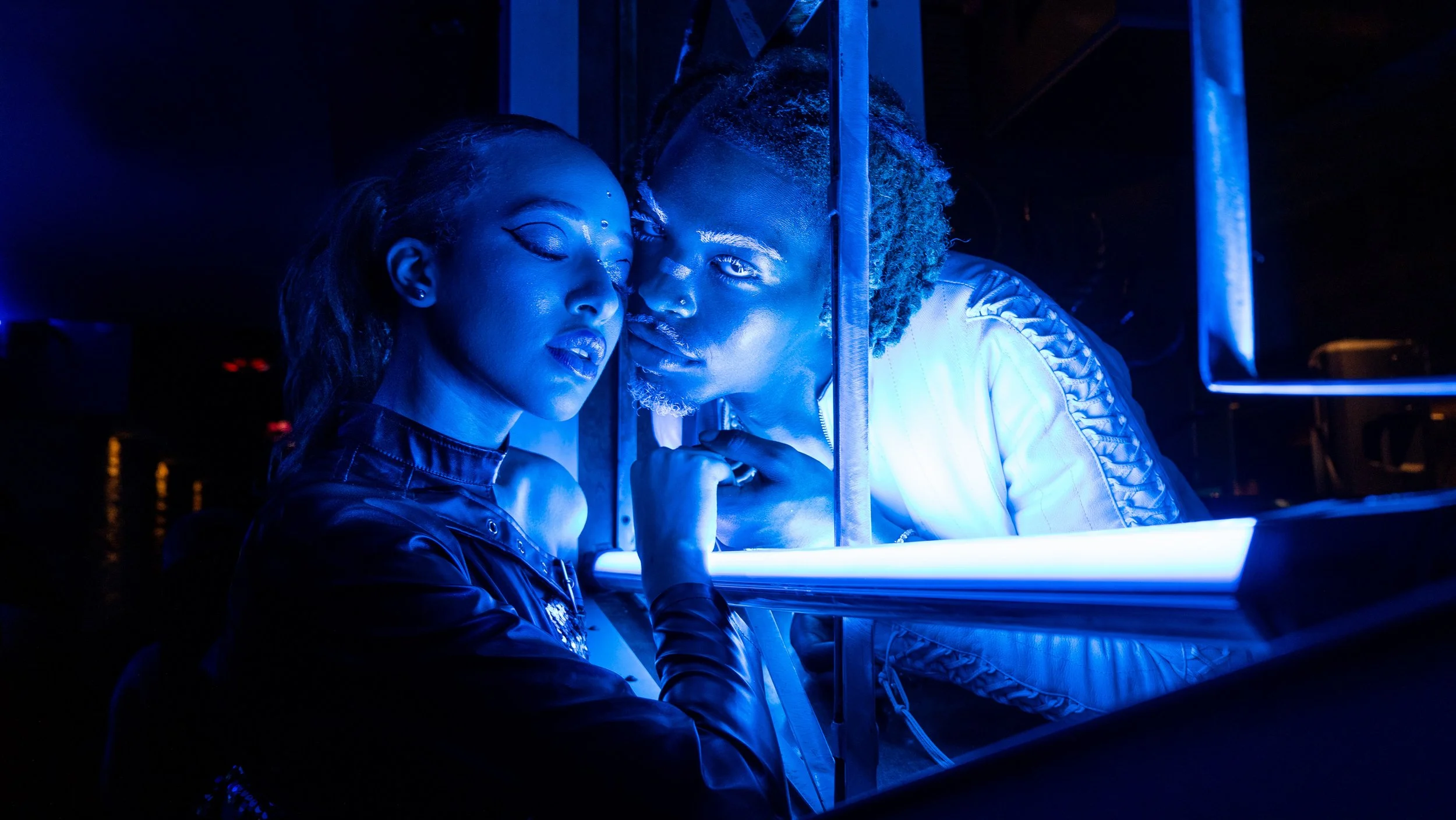 Two individuals with dark skin leaning close to each other in a dark room illuminated by blue light, with a metal bar between them.