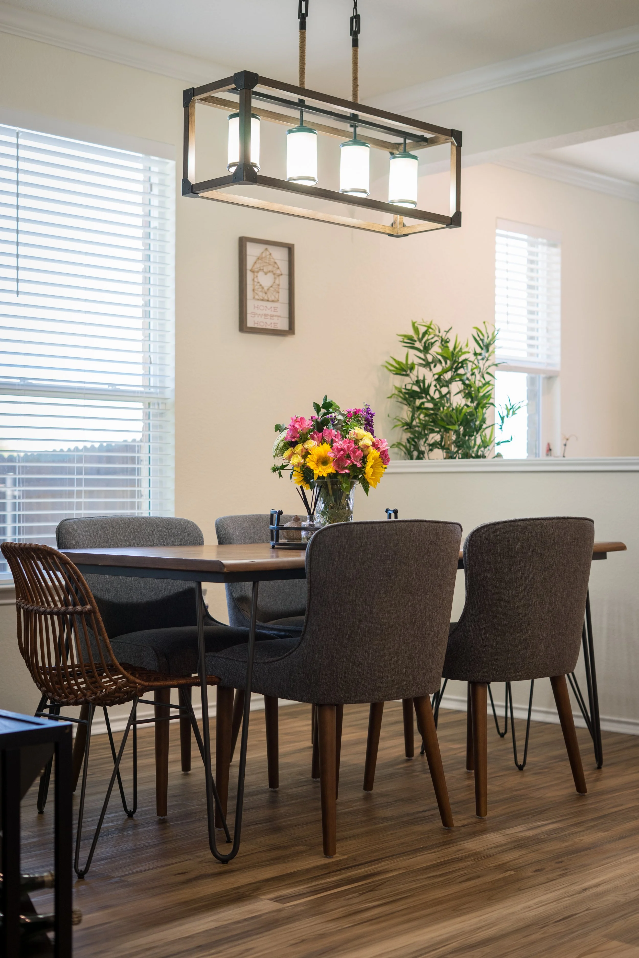 A dining room with a wooden table, surrounded by six chairs, and a bouquet of colorful flowers in the center. There is a modern light fixture hanging overhead and two windows with blinds in the background, along with a small framed wall art.