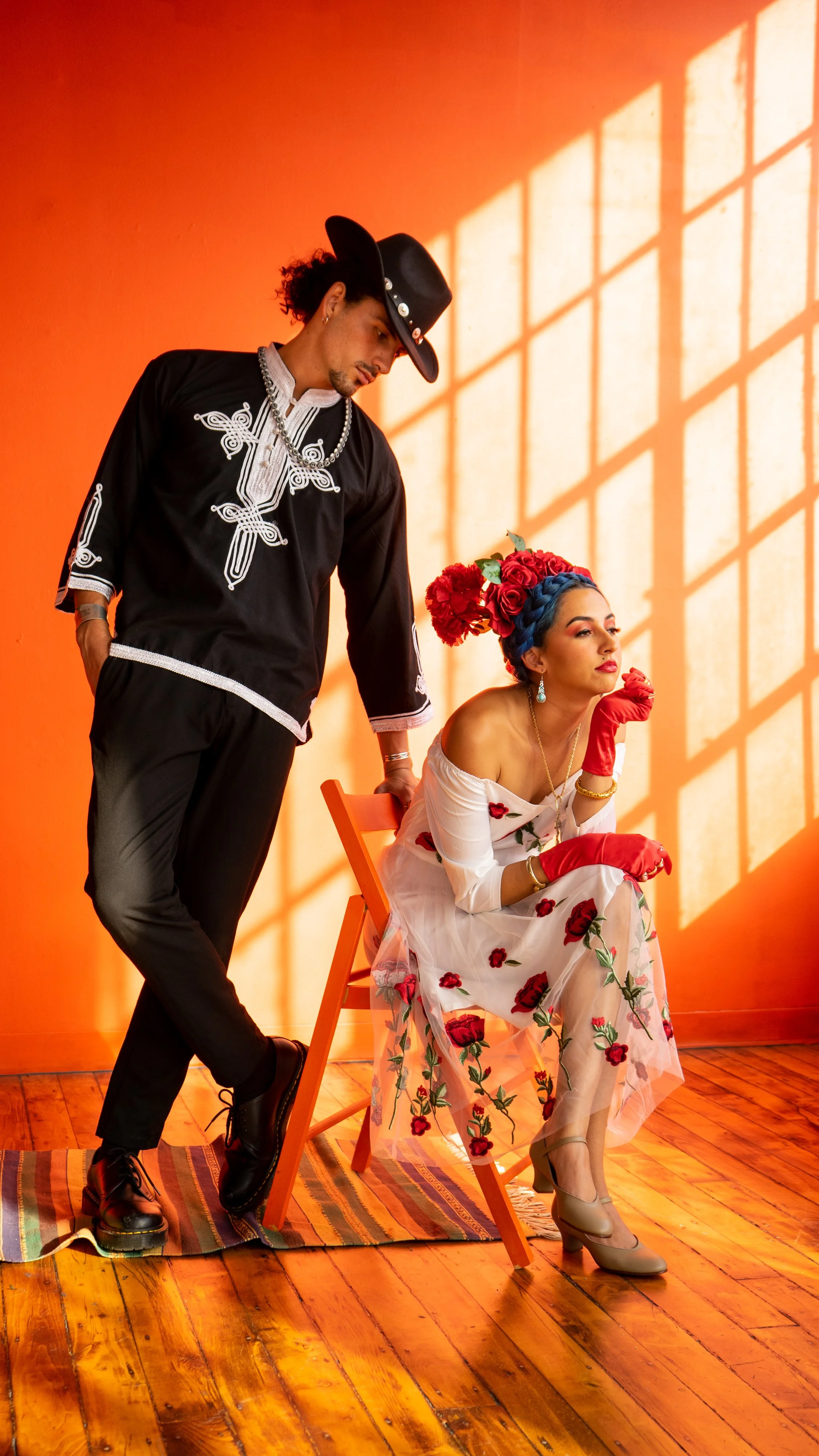 A woman with blue hair and a floral dress sits on a chair, wearing red gloves, necklace, earrings, and a gold bracelet, while a man in traditional Mexican charro attire stands beside her against an orange wall with a window shadow pattern.