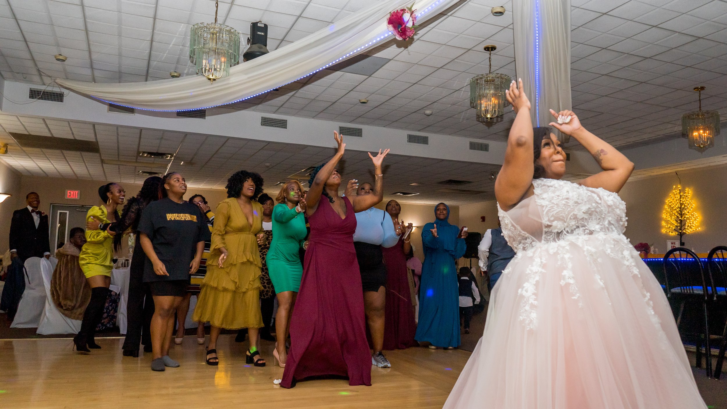 A bride in a white wedding gown with floral lace details prepares her bouquet during her wedding reception, while a group of women and a man on the dance floor watch and cheer.