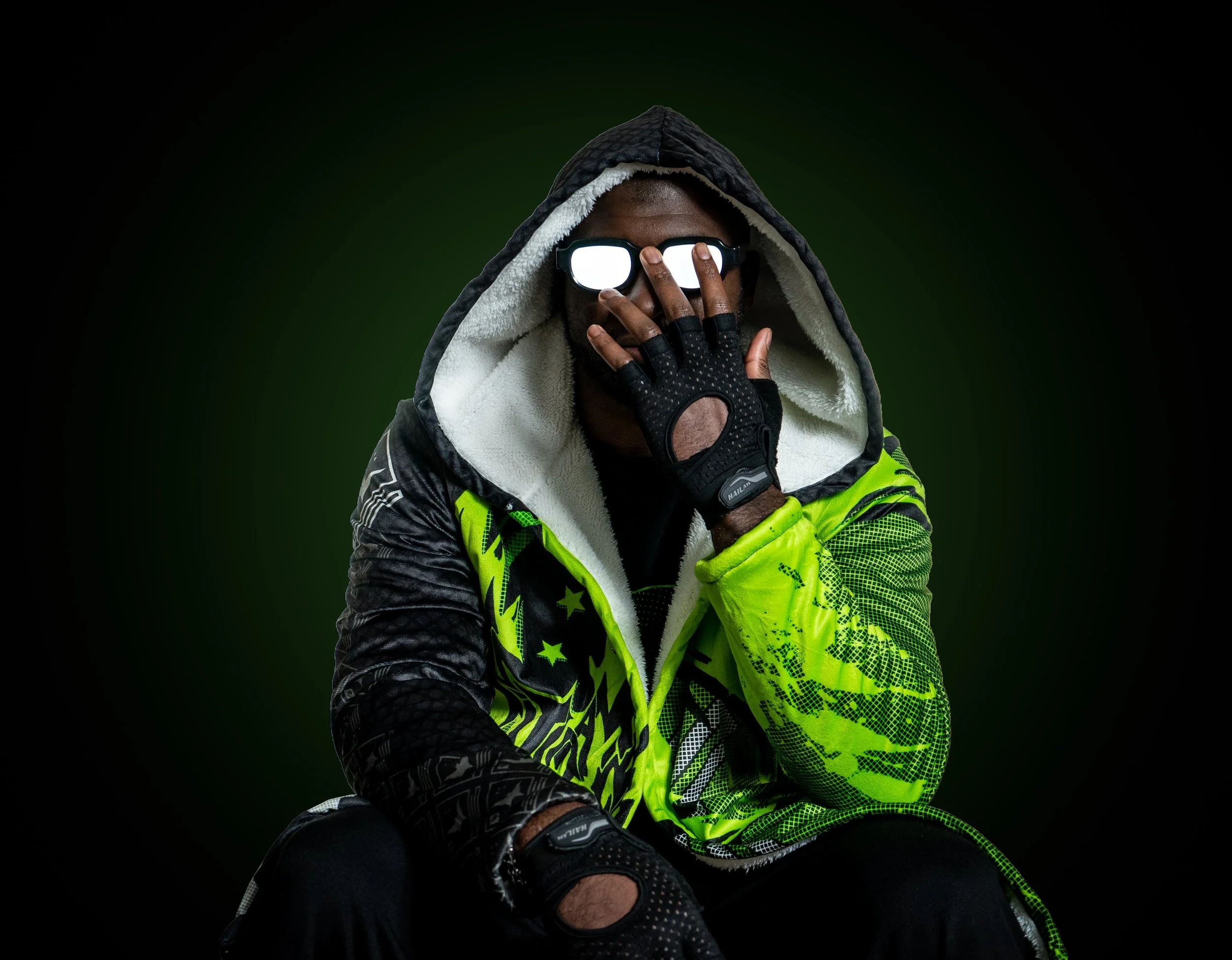 A person wearing a neon green and black jacket with a hood, black gloves, and sunglasses, sitting against a dark background.