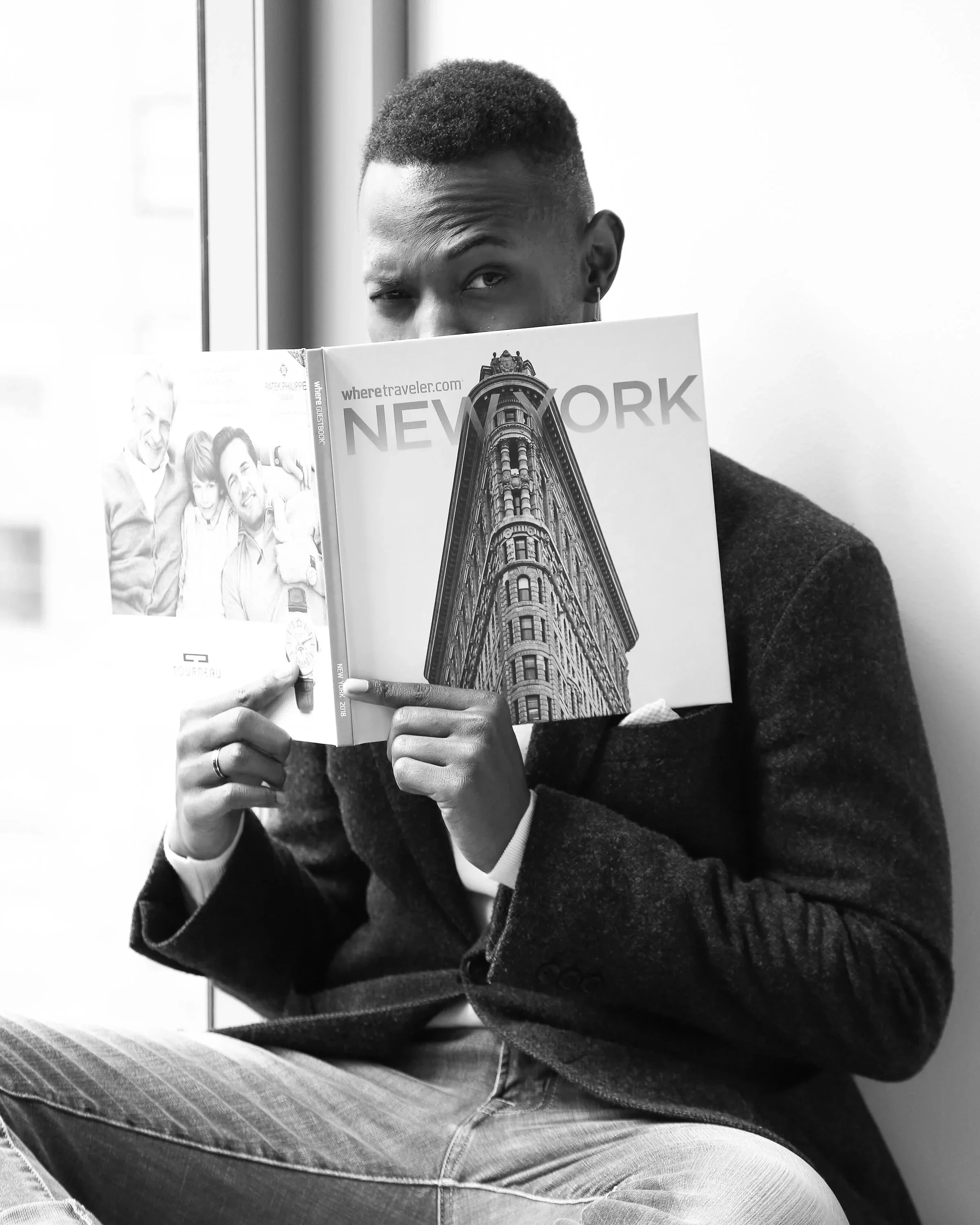A person with short hair and wearing a dark jacket and jeans sitting in front of a window, holding a magazine with the cover showing a tall building in New York City, partially hiding their face.