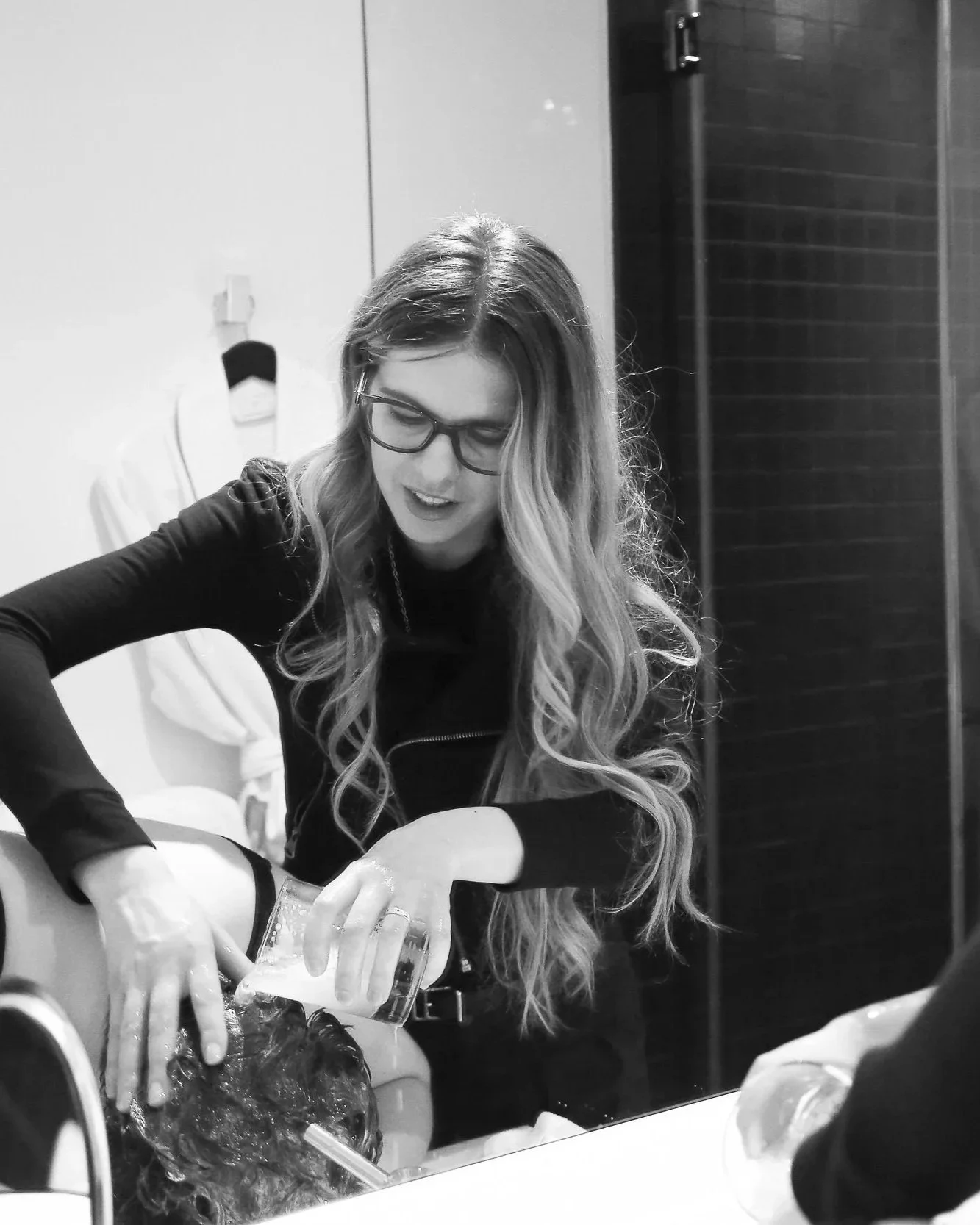 A woman with glasses and long wavy hair pouring a cup of water over a person's head.