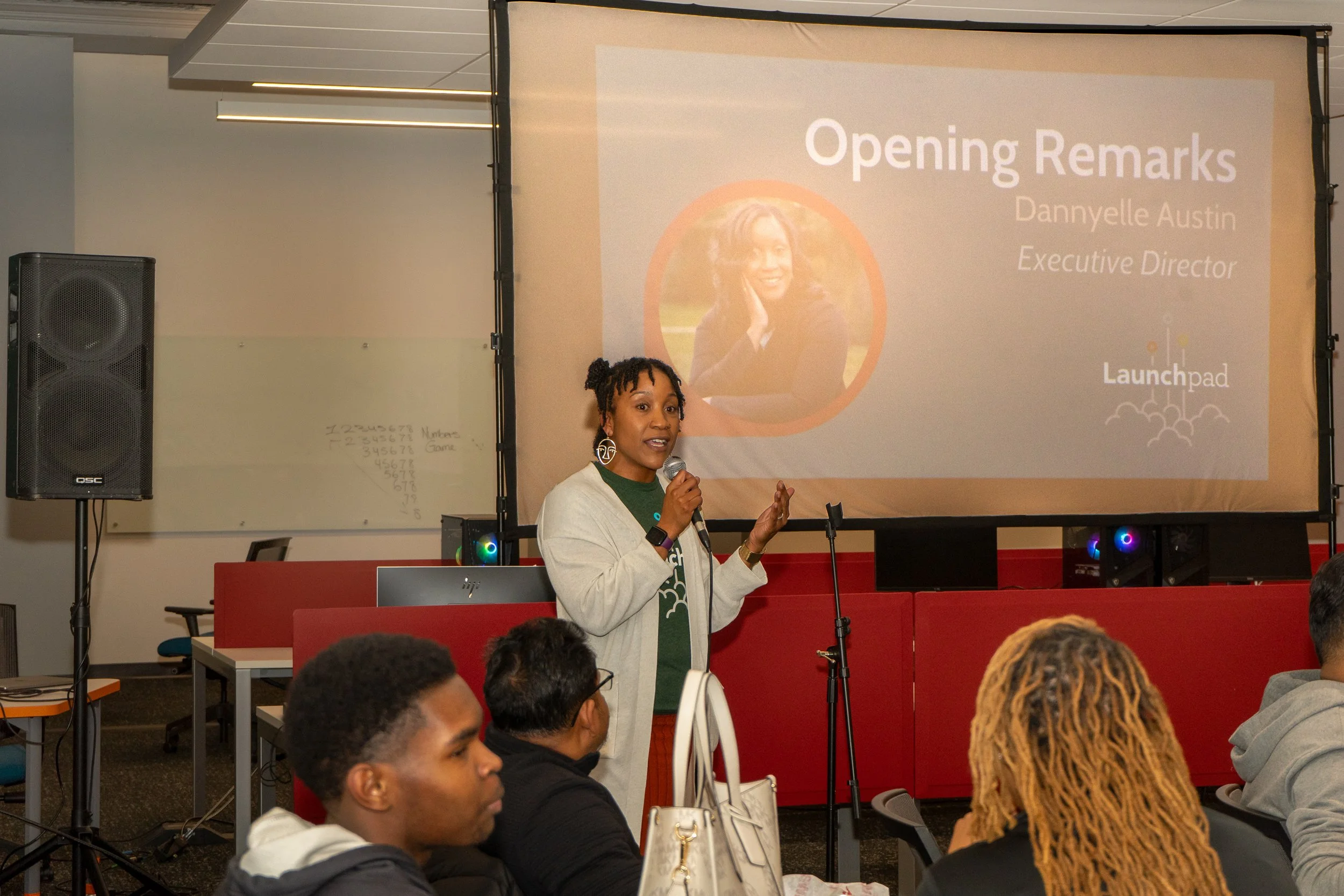 Woman giving a presentation with a microphone in front of a projected slide that reads 'Opening Remarks Dannyle Austin, Executive Director' at Launchpad conference.