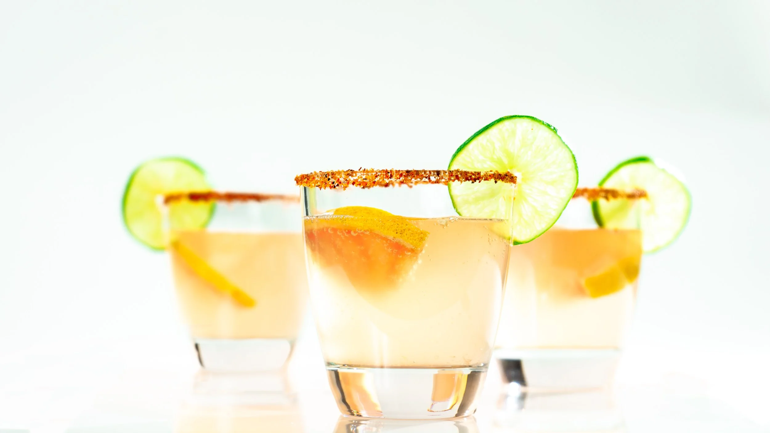 Three glasses of citrus cocktail with lime slices and a salted rim, arranged in a line on a white background.