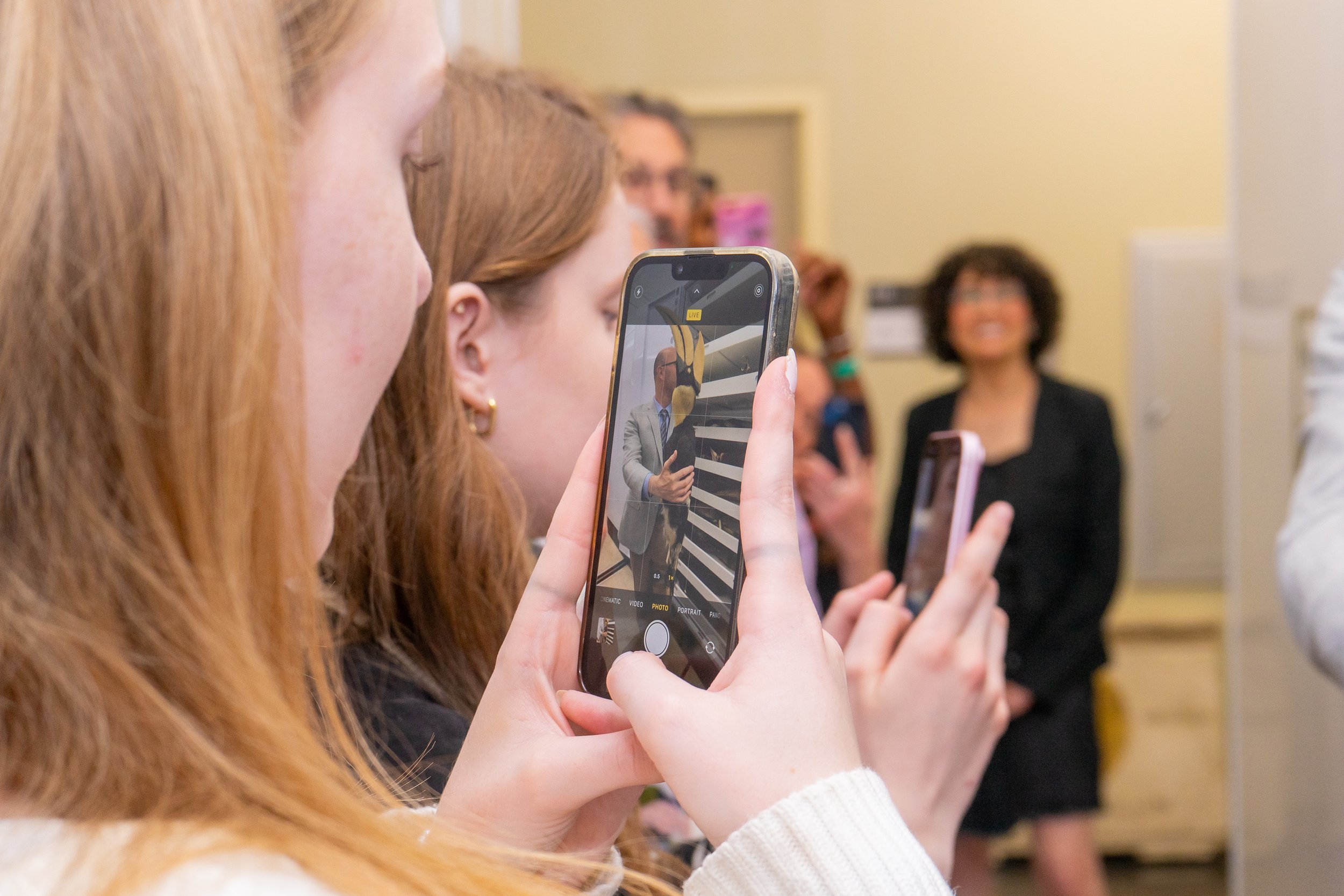 A group of people, mostly women, taking photos with smartphones at an indoor event, with a woman in a black blazer smiling in the background.