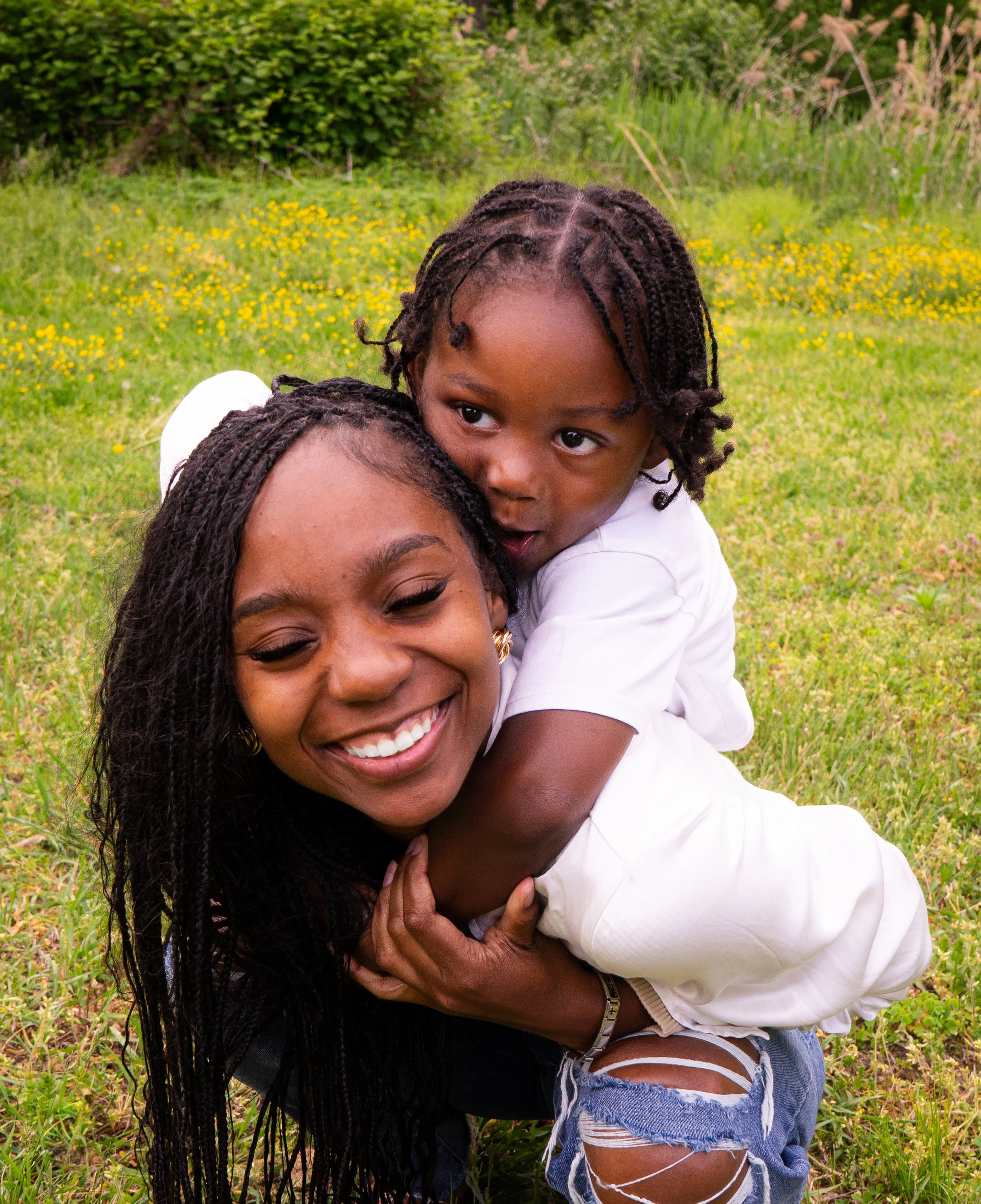 A joyful woman and child playing outdoors on a grassy field with yellow flowers, a woman with long dark hair and a child with short curly hair, both smiling.