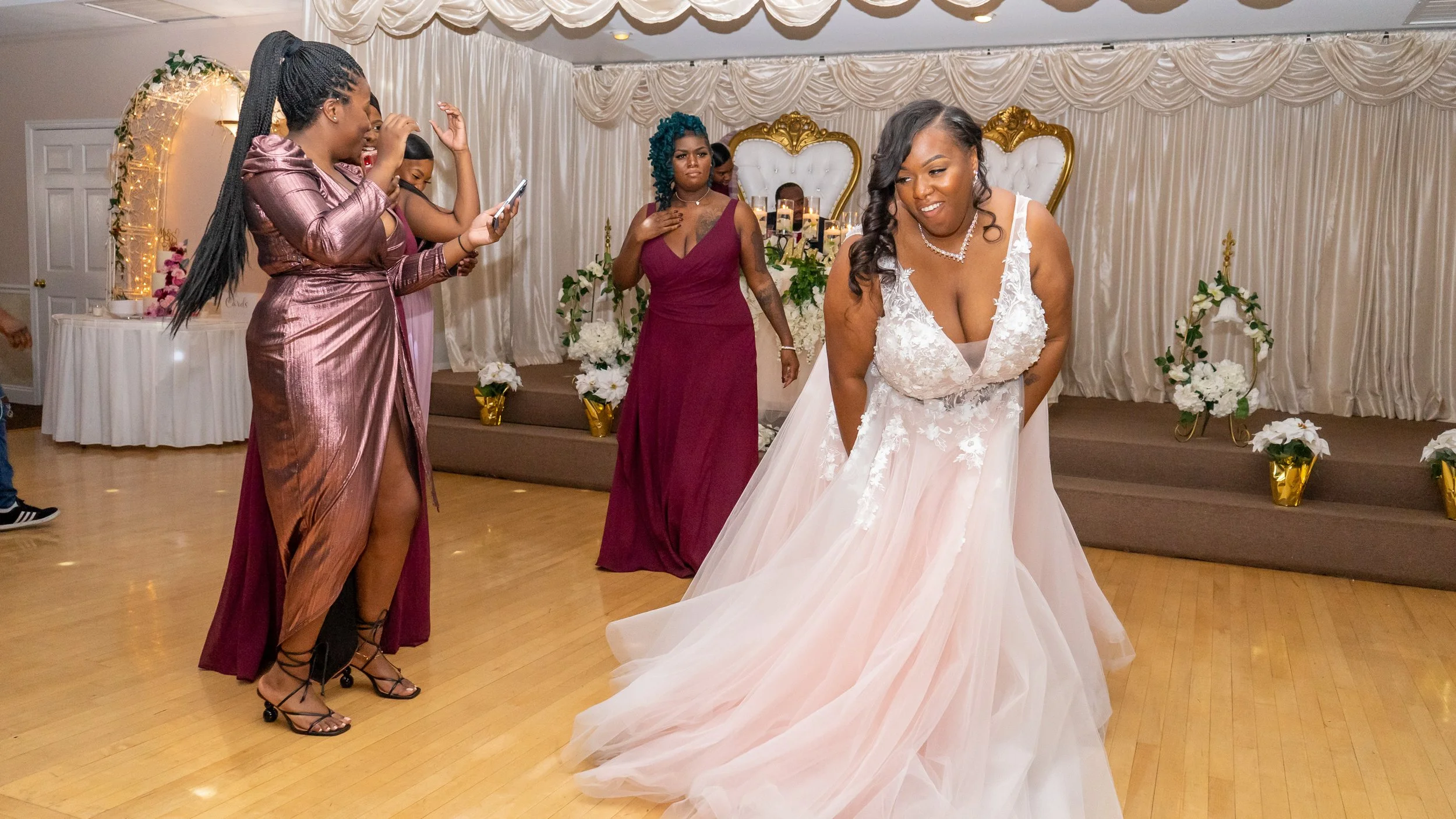A bride in a white wedding gown dancing with a smile at a wedding reception, surrounded by guests in formal attire, with draped curtains, floral decorations, and gold accents in the background.