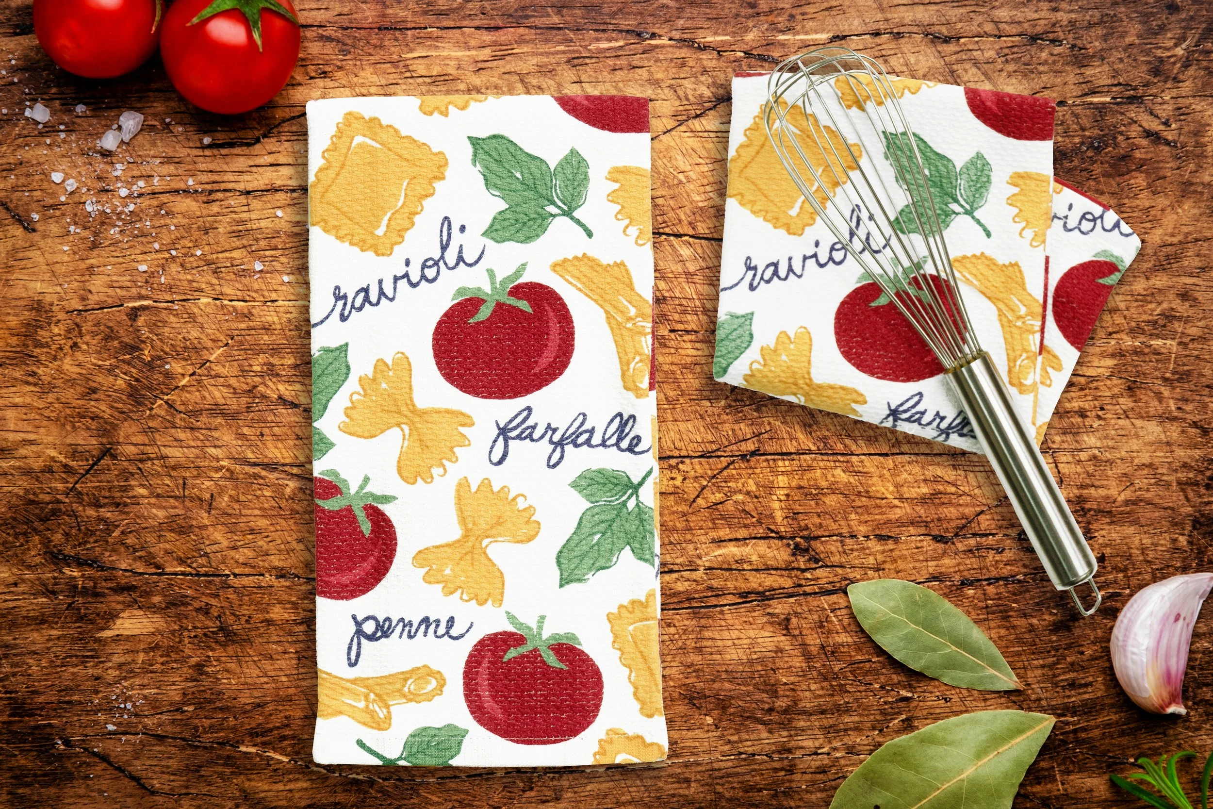 Kitchen towel with tomato, basil, pasta, and ravioli pattern placed on a wooden surface with tomatoes, garlic, and bay leaves nearby, and a wire whisk on the towel.