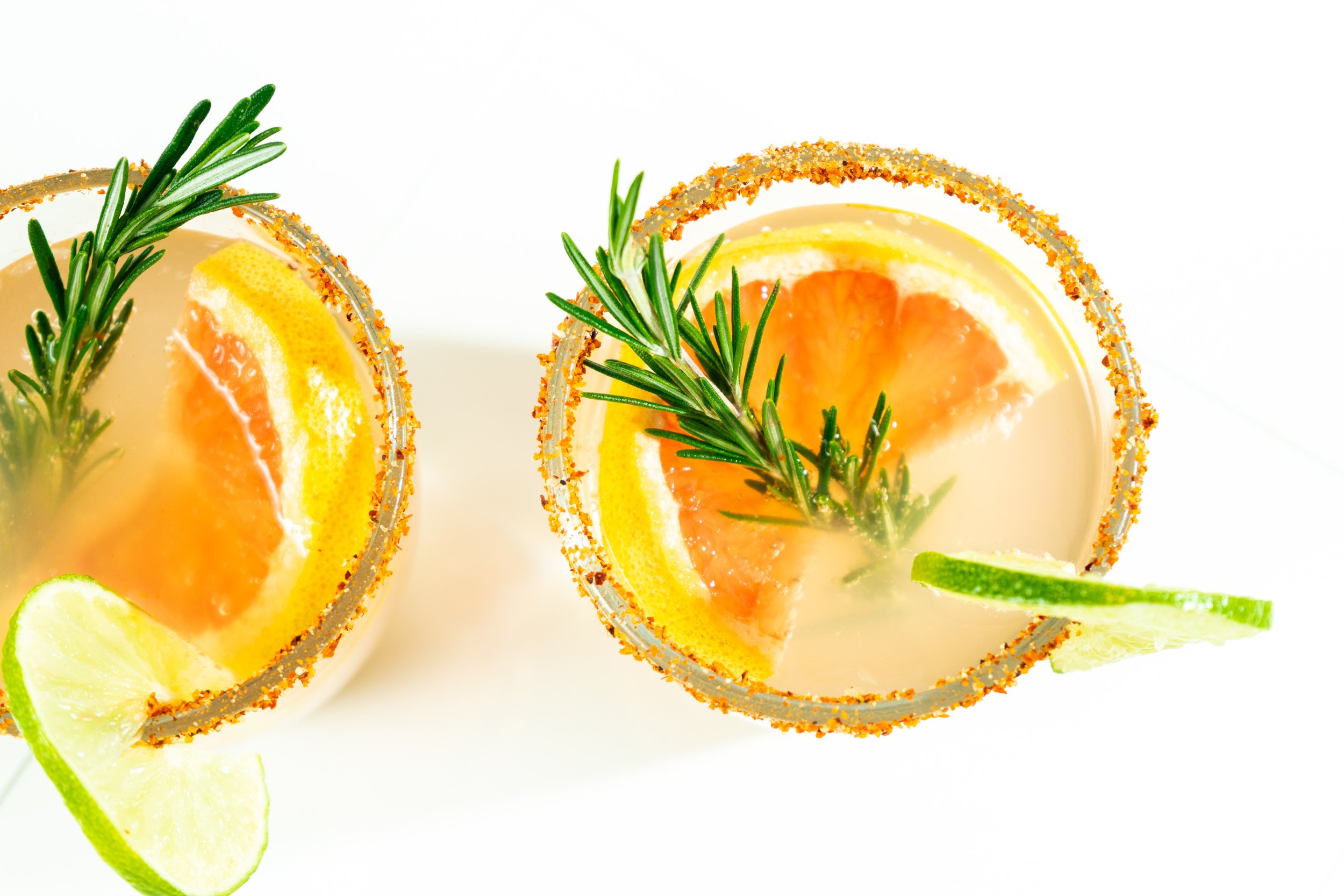 Two cocktail glasses rimmed with salt, garnished with lime and orange slices and sprigs of rosemary on a white background.