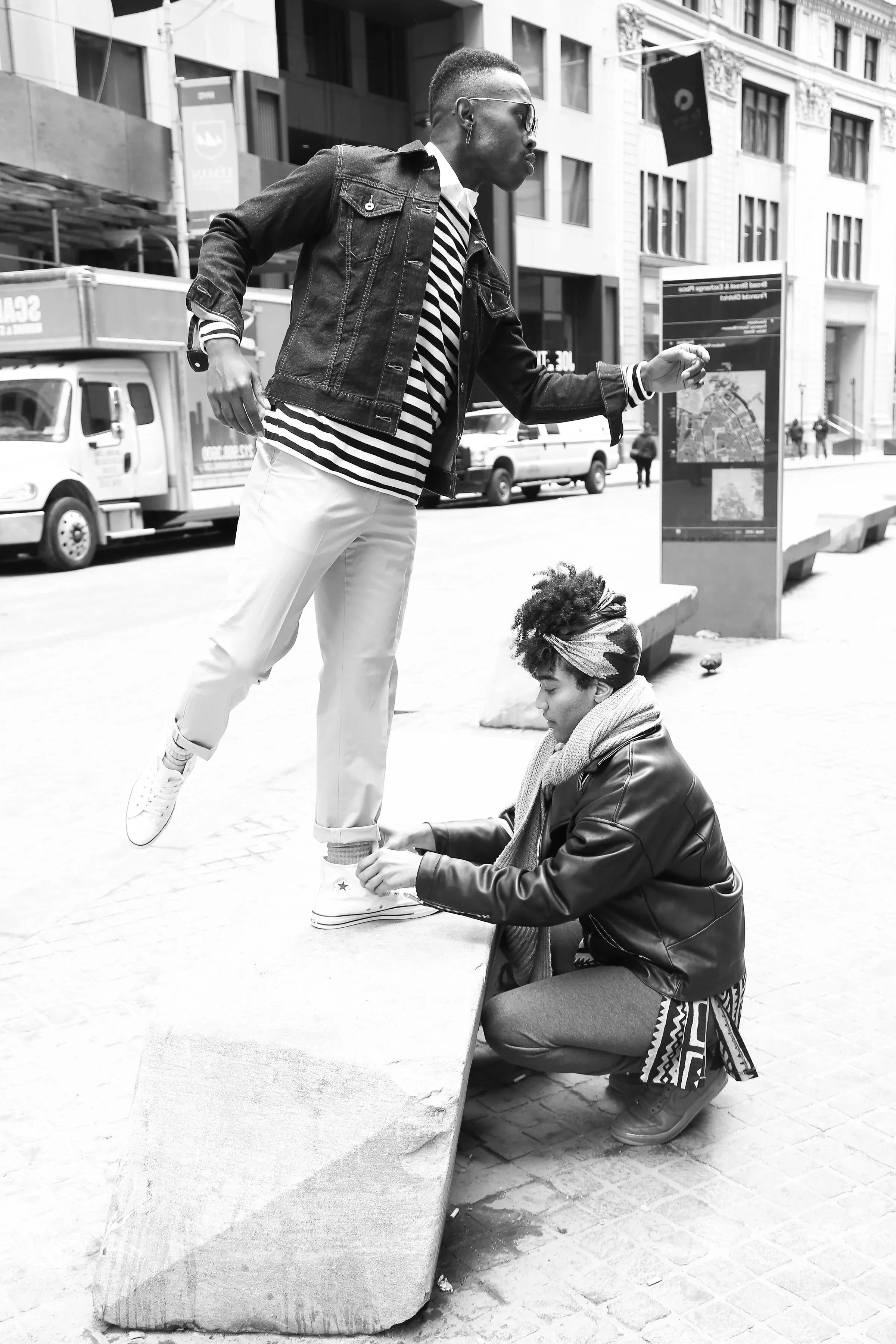 A man appears to be levitating above the ground while a woman secures his shoe on a city sidewalk. The photo is black and white.
