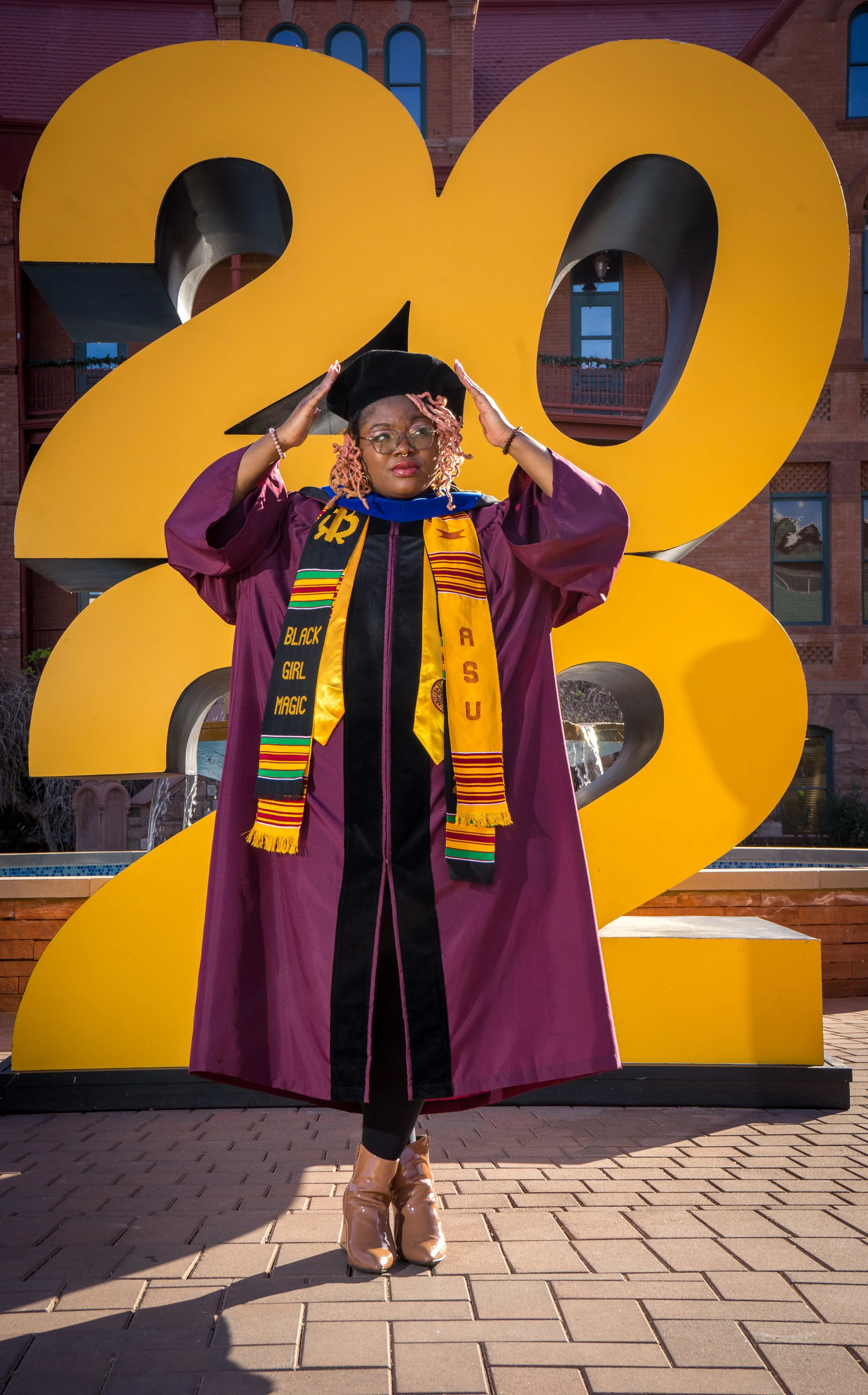 A woman in graduation regalia standing in front of a large yellow '2023' sculpture, adjusting her graduation cap.