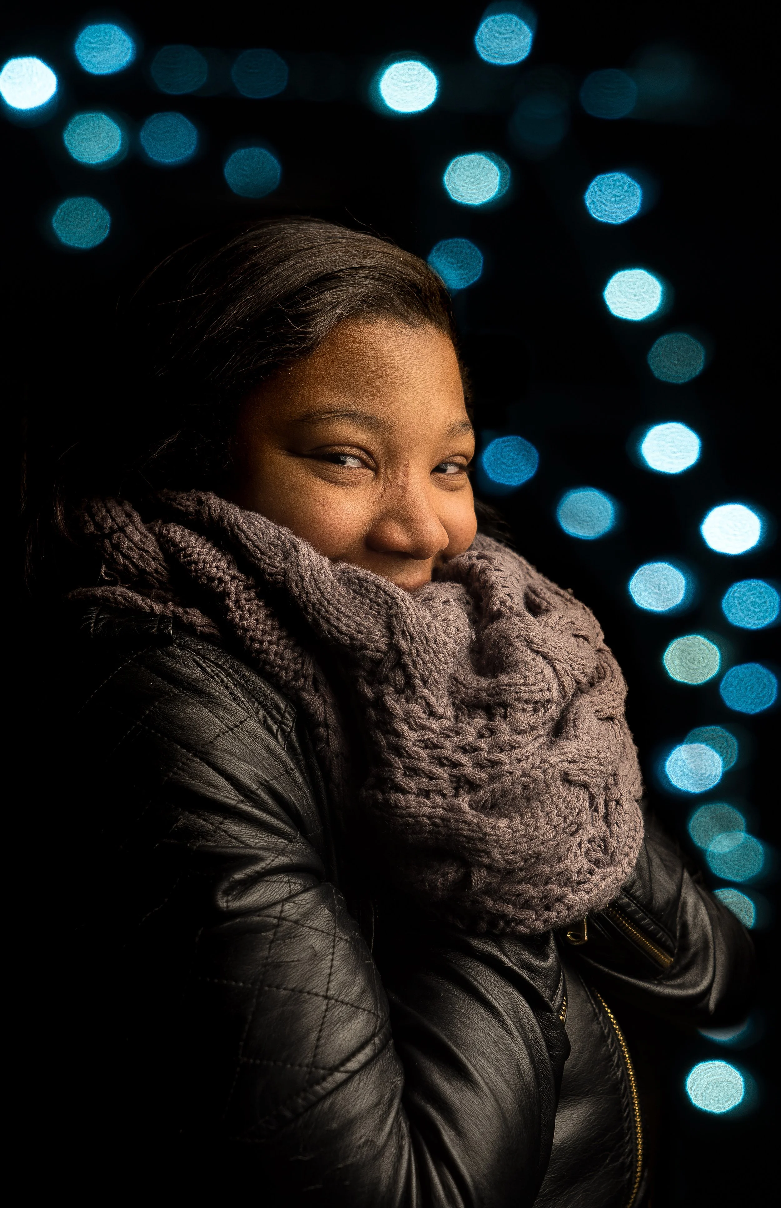 Young woman with dark hair smiling, wearing a gray knit scarf and black leather jacket, at night with blurred blue and white bokeh lights in the background.