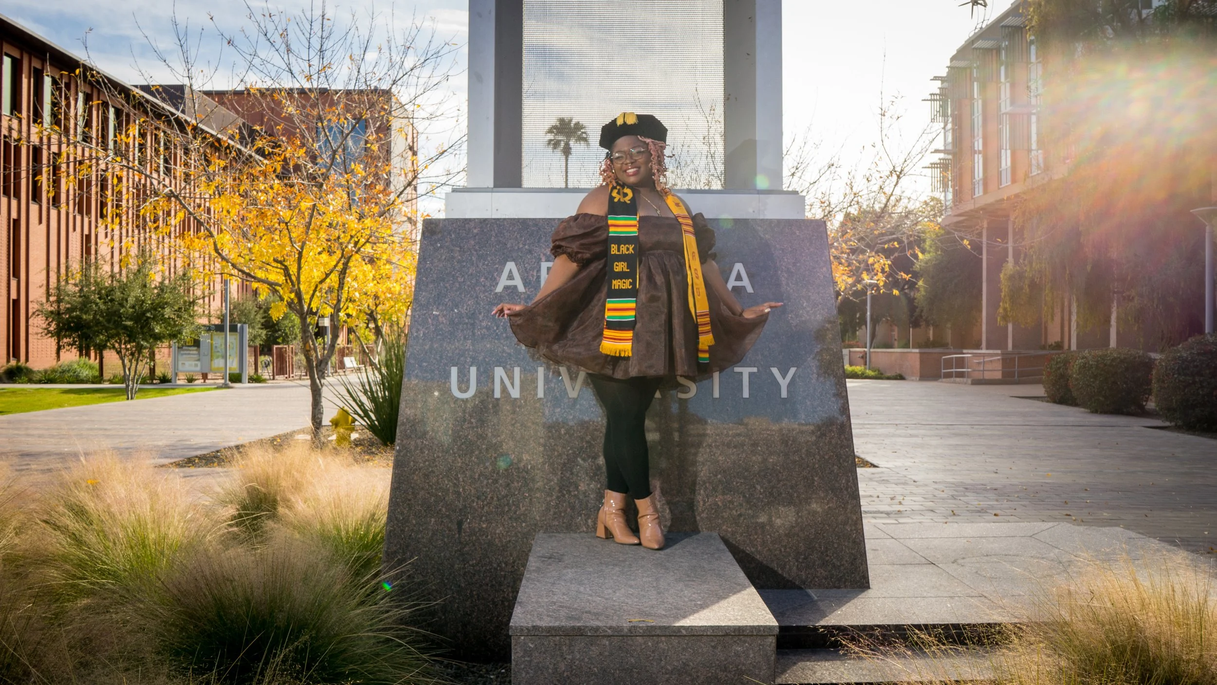 A woman celebrating graduation on a university campus, wearing a cap and gown with an Afrocentric stole, standing in front of a university sign with trees and buildings in the background and autumn foliage.