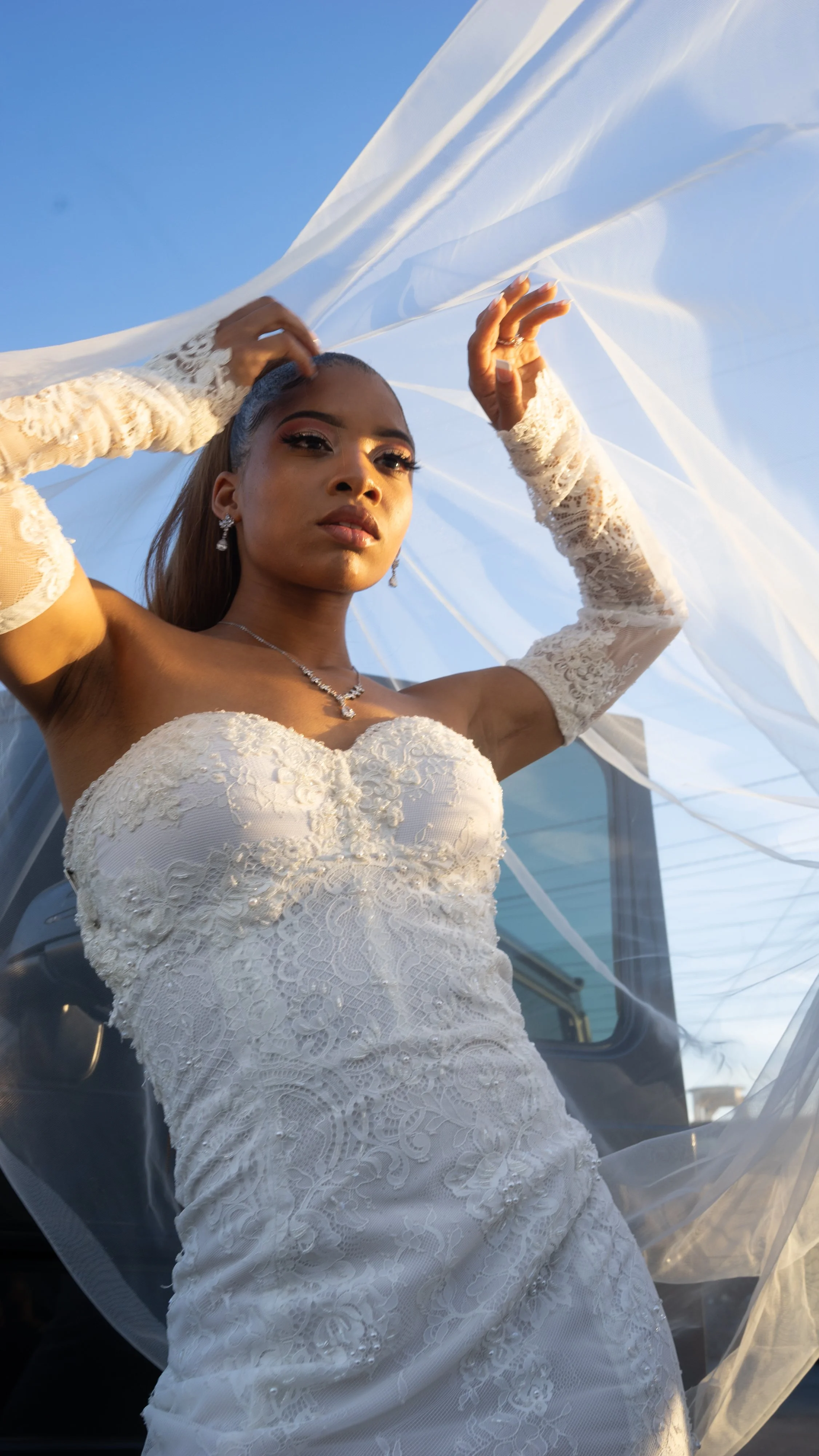A woman in a strapless white lace wedding dress with lace gloves, jewelry, and makeup, standing outdoors with blue sky in the background, holding a sheer fabric above her head.