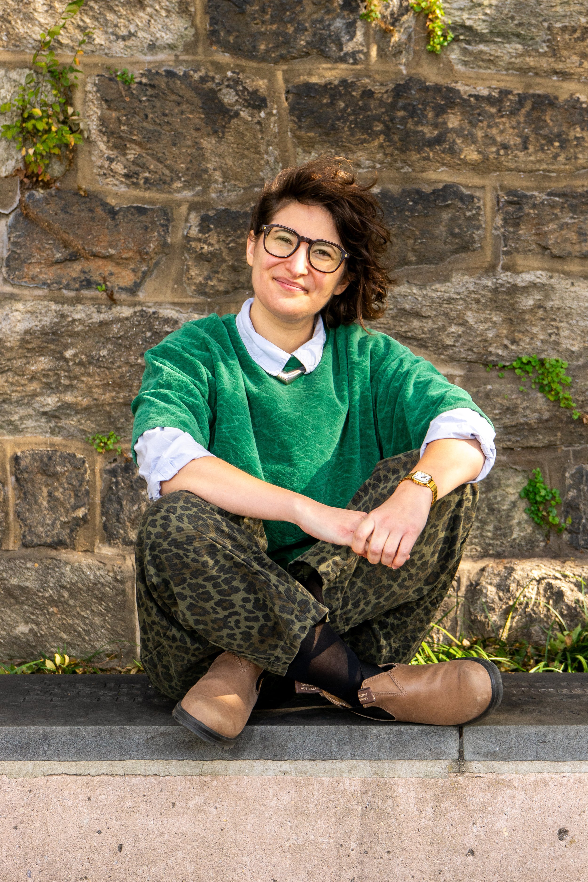 A woman with curly brown hair and glasses sitting cross-legged on a ledge, wearing a green shirt, leopard print pants, and tan boots, smiling in front of a stone wall with small green plants.