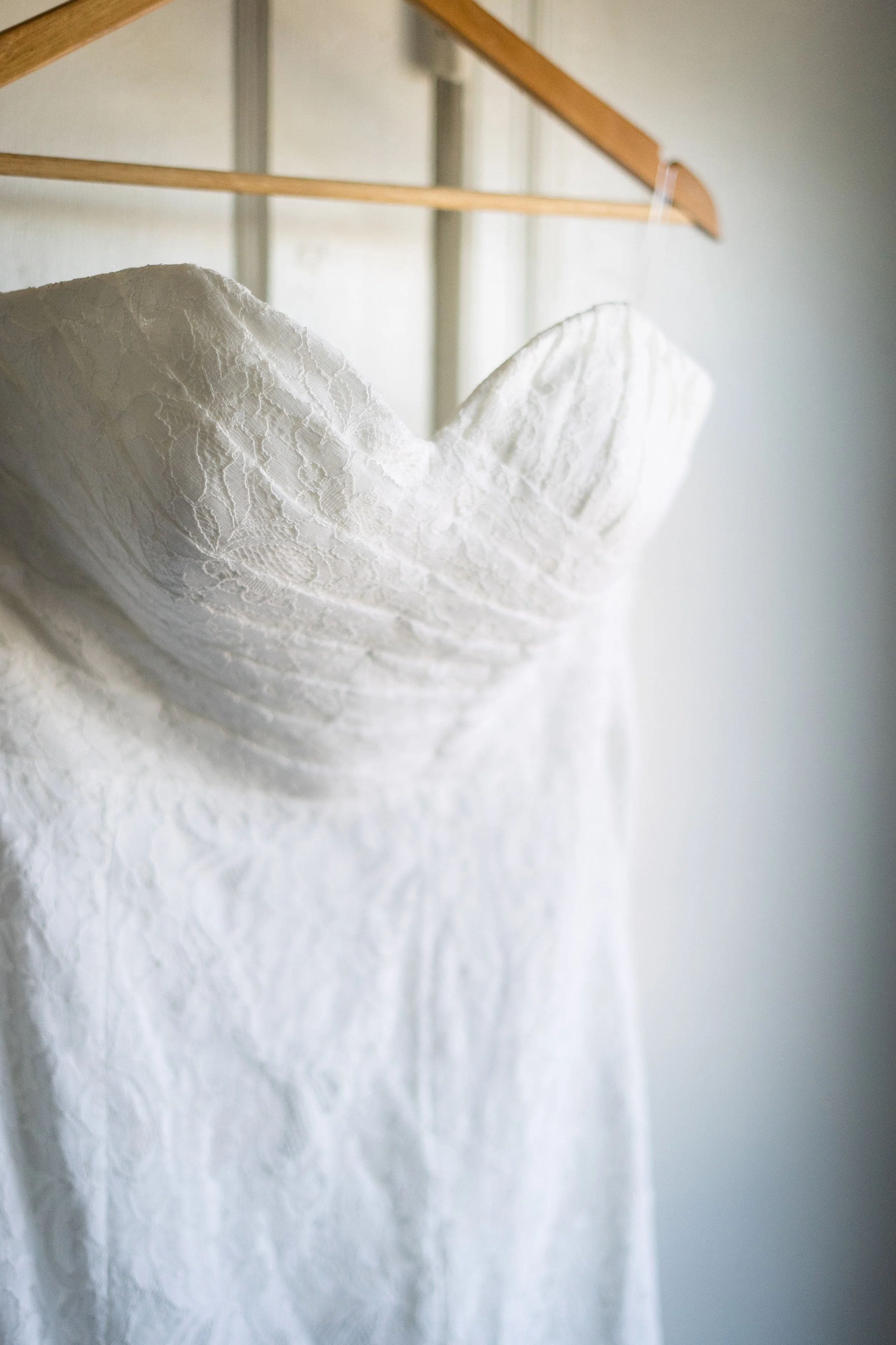 A white wedding dress with lace fabric hanging on a wooden hanger against a neutral wall.