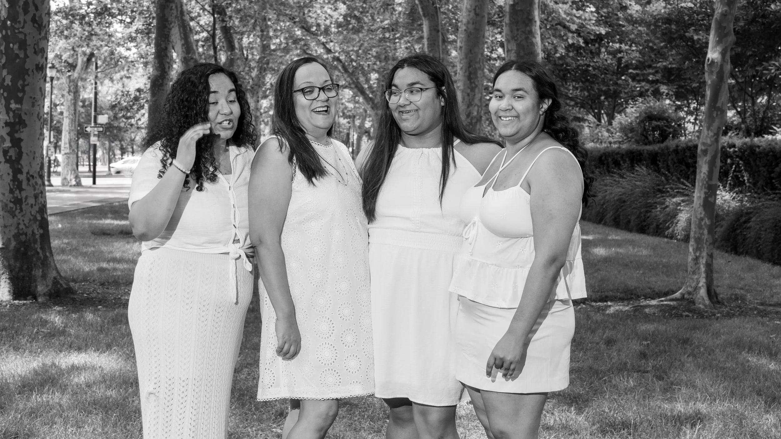 Four women standing together outdoors in a park, smiling and talking amidst trees and grass.