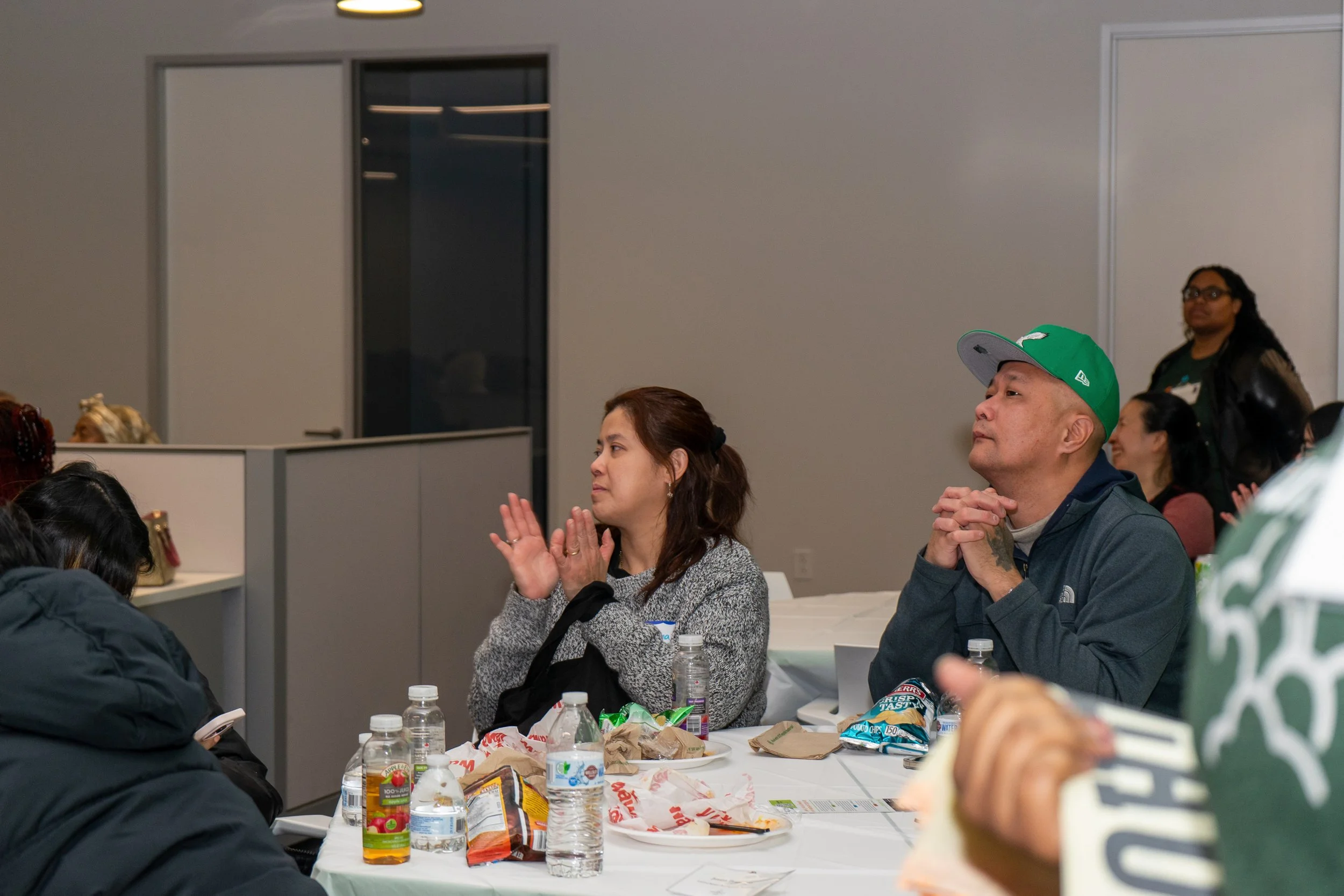 People attending a meeting or event, seated around a table with snacks and drinks, some with attentive or thoughtful expressions.