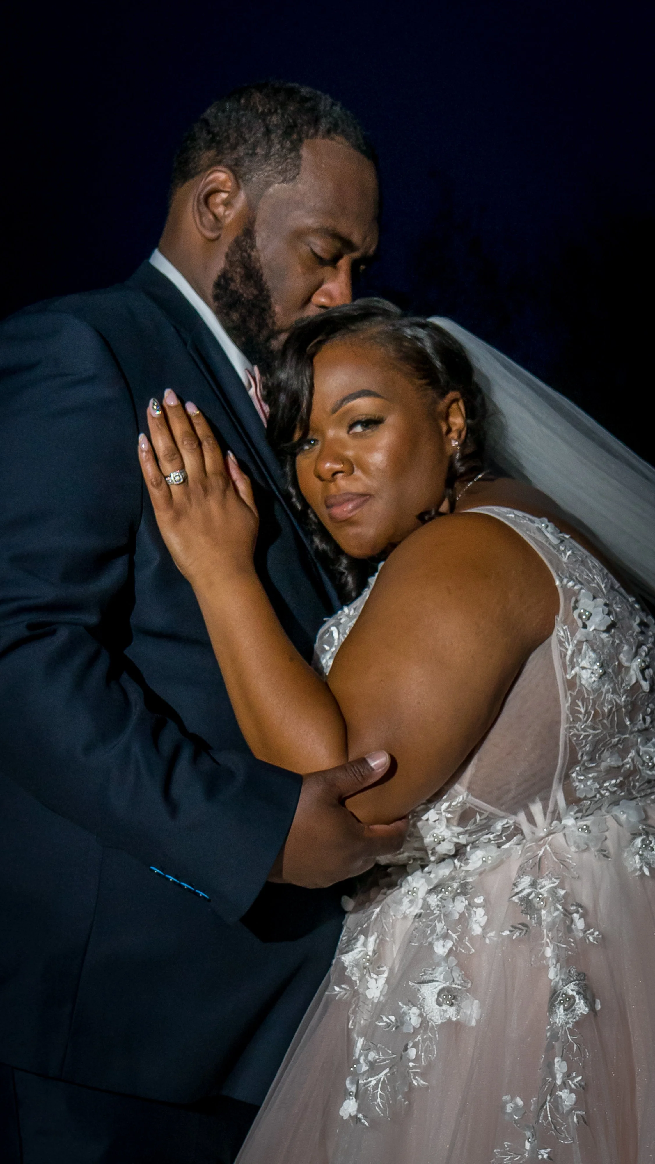 A newlywed couple embraces closely, with the groom wearing a dark suit and the bride in a white wedding gown with floral embroidery, against a dark background.