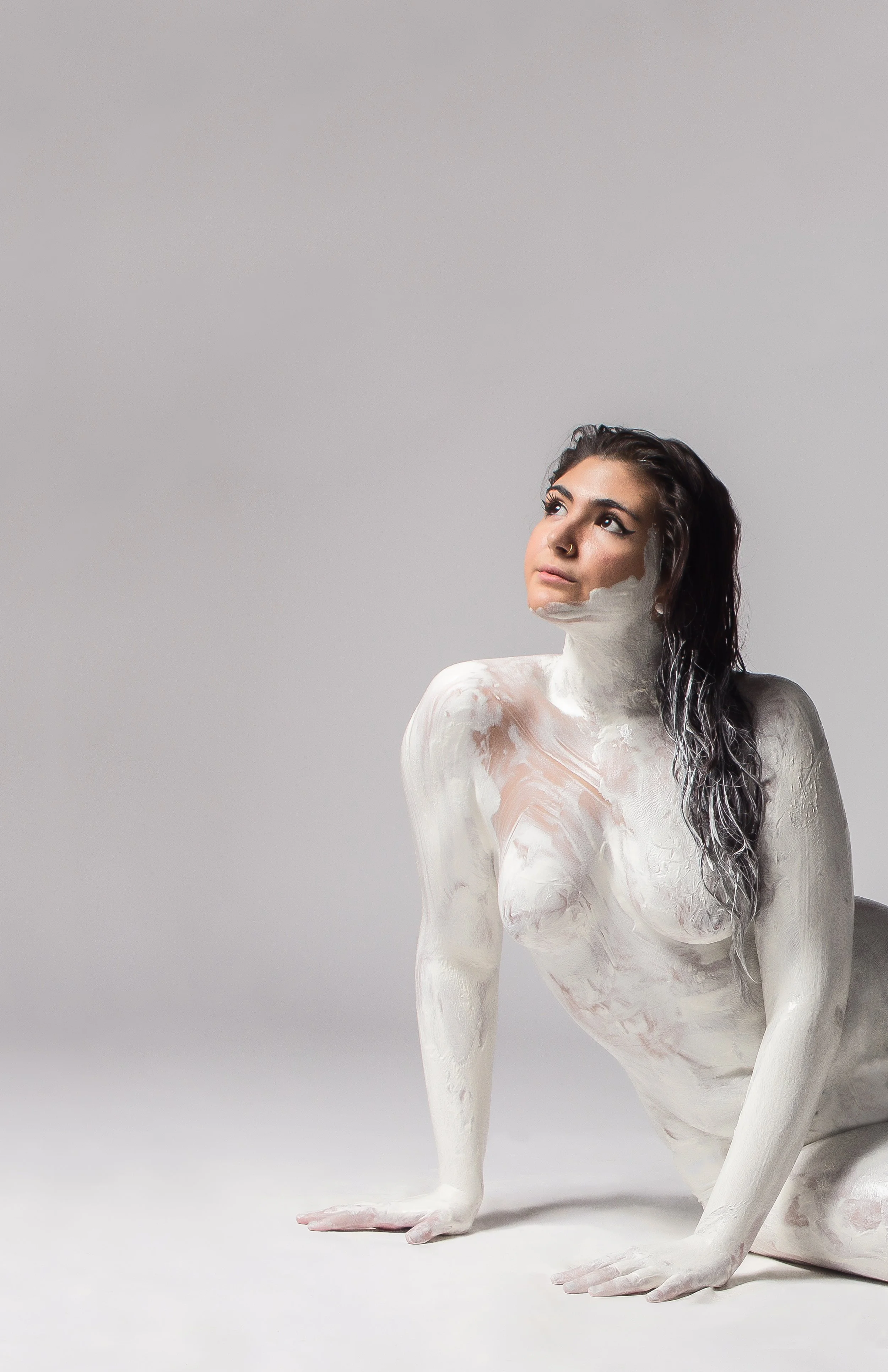 A woman with dark, wet hair posed on her hands and knees, with her body covered in white paint, against a plain gray background.