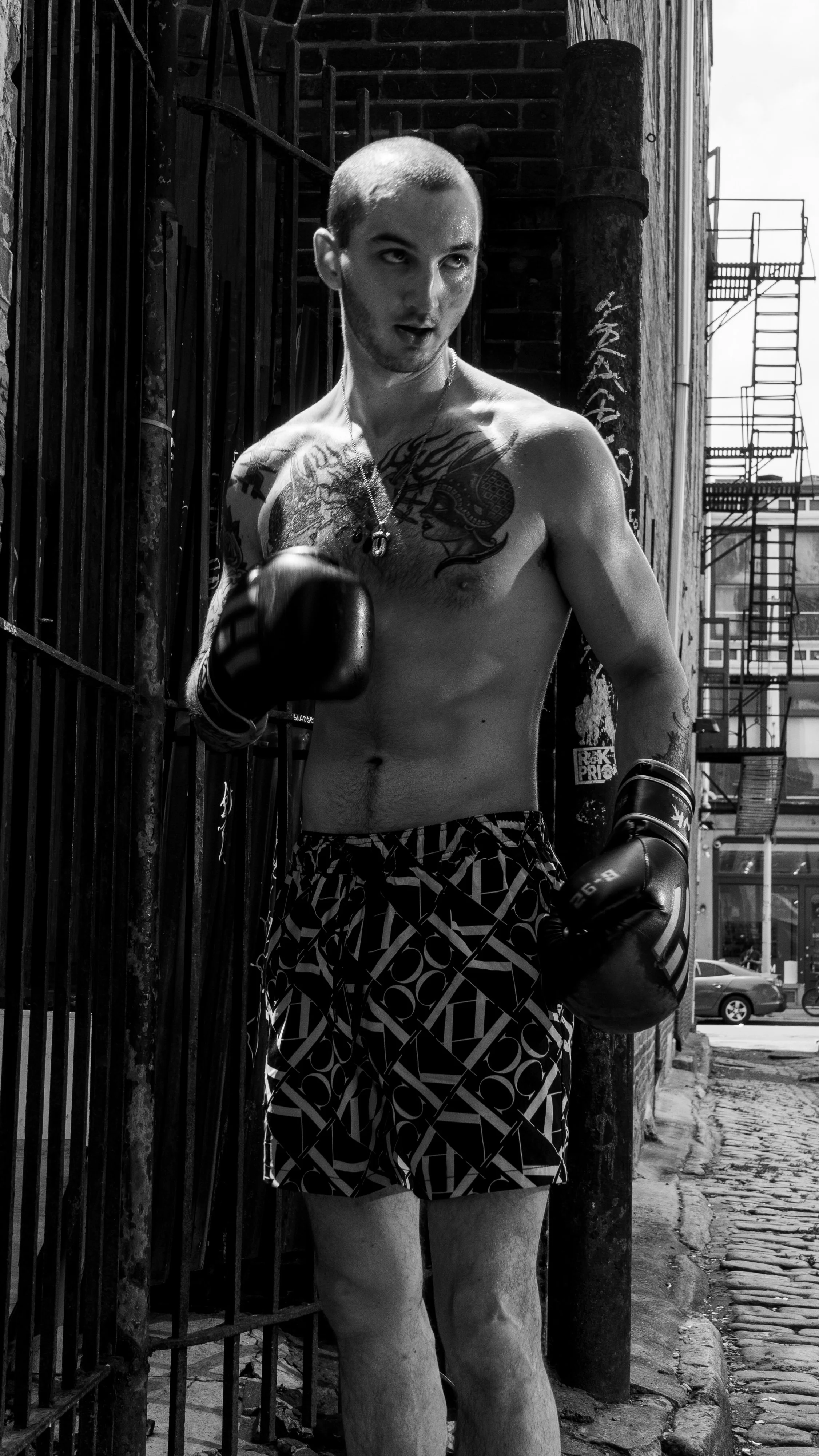 A shirtless man with tattoos wearing boxing gloves and patterned shorts, standing outdoors next to a metal fence and brick wall, with city buildings and cars in the background.