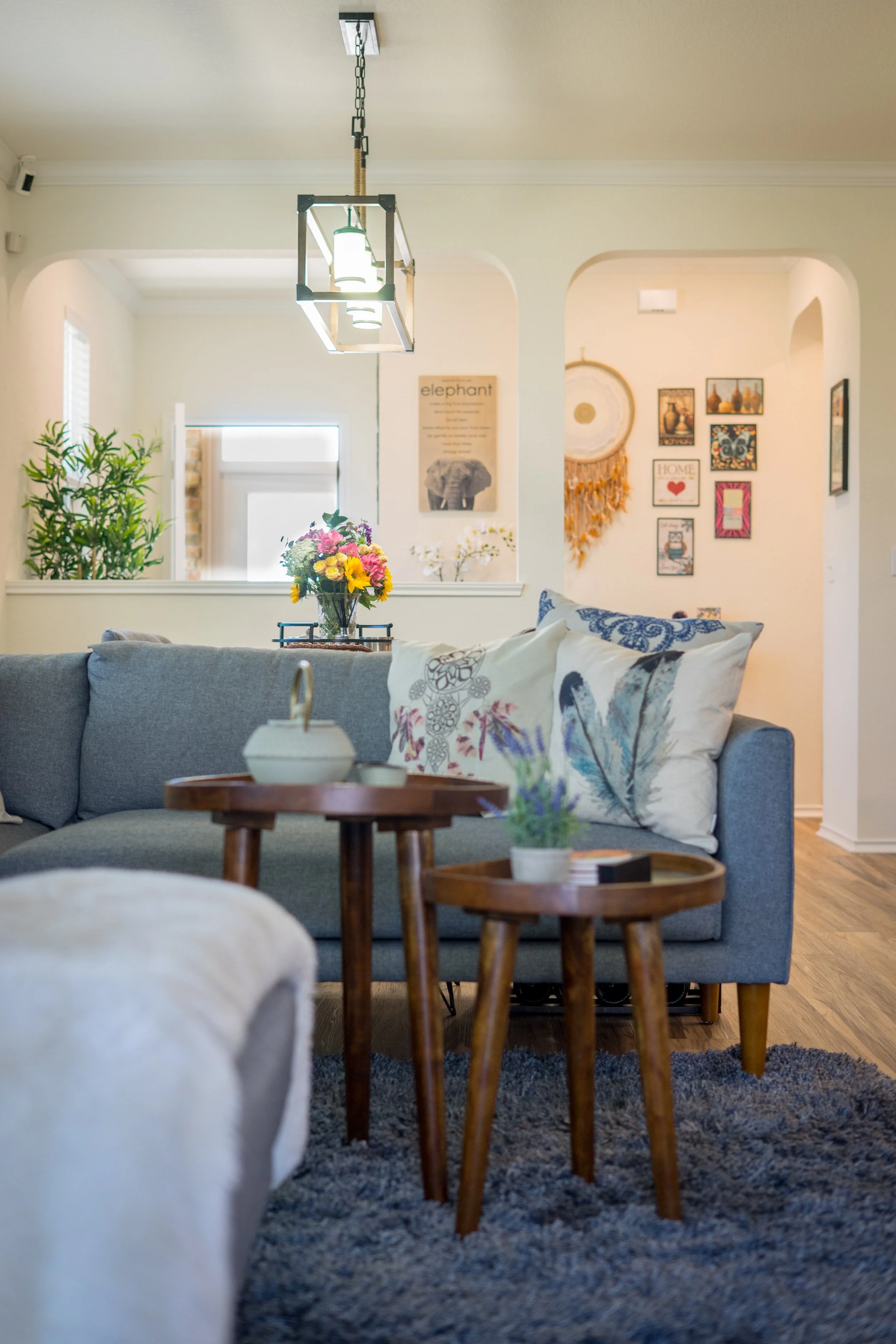 Living room with gray sofa, decorative pillows, small wooden tables with potted plants and a white teapot, wall art, and a hanging light fixture.