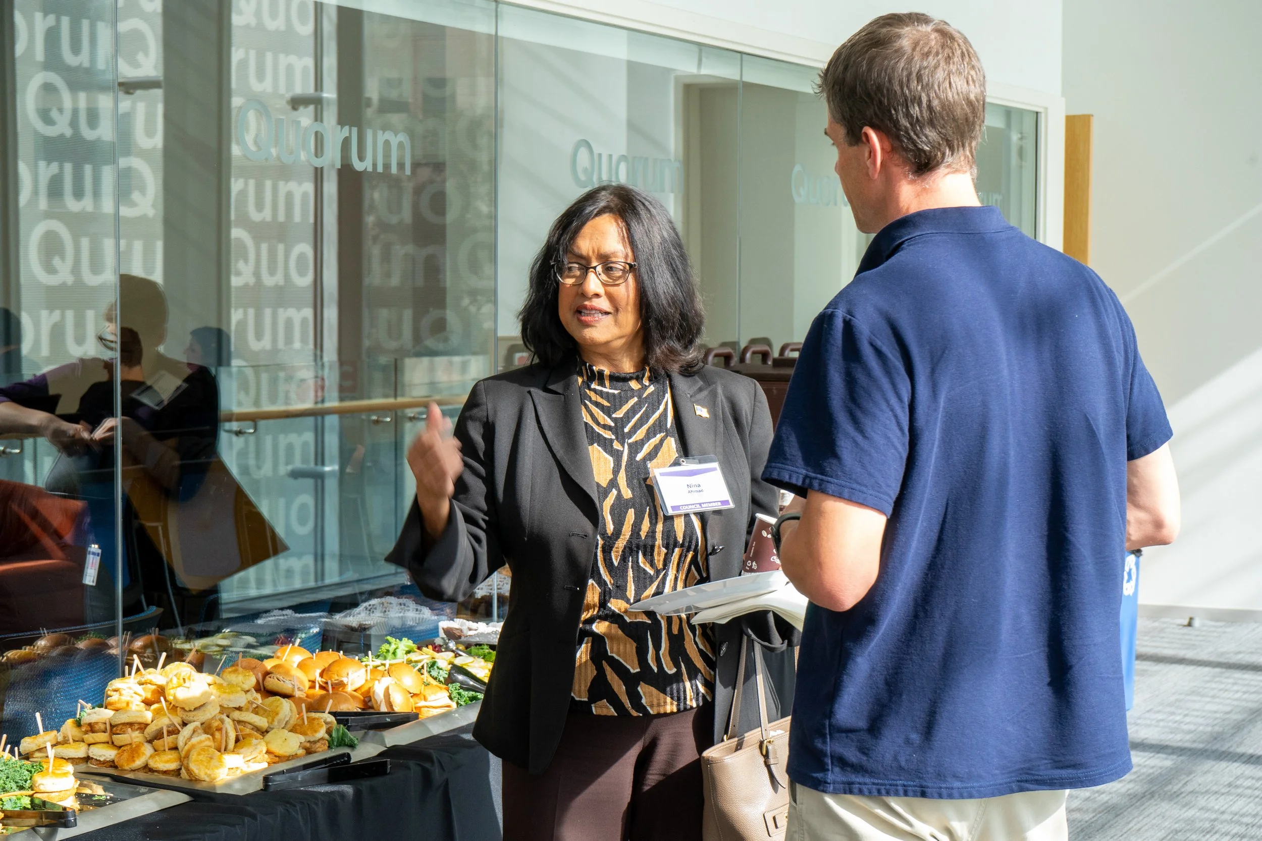 A woman and a man having a conversation at a breakfast buffet table during a conference or meeting, with trays of sandwiches and snacks visible.