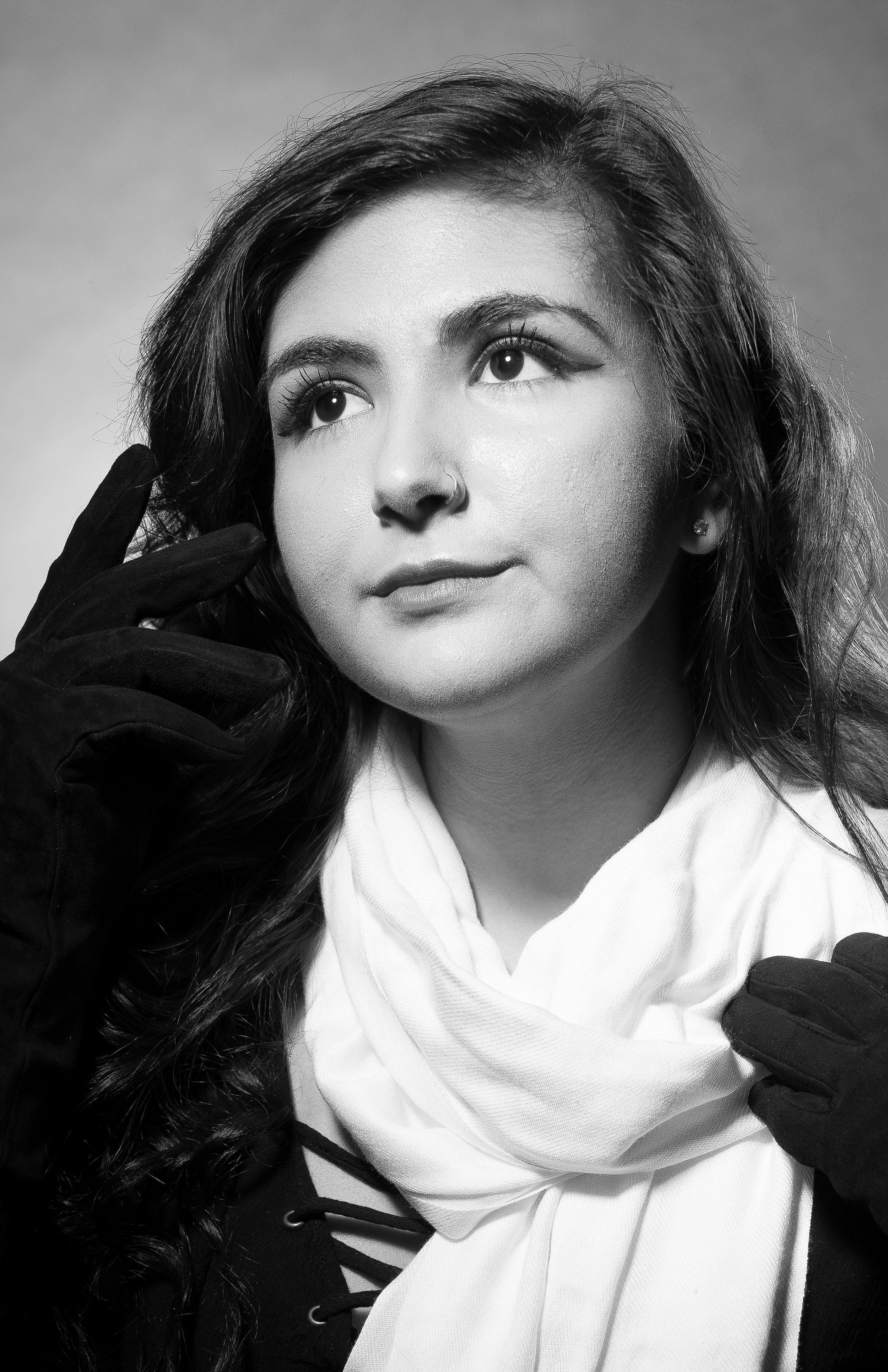 Black and white portrait of a young woman with wavy hair, wearing gloves, a white scarf, and a top with lace-up detail.