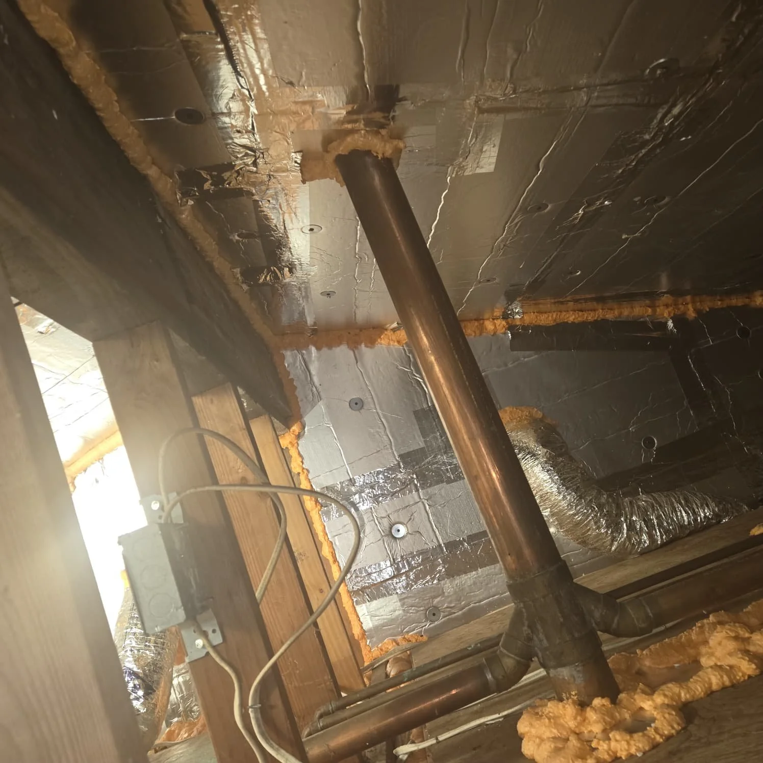 Indoor construction scene showing copper pipes, insulation foam, and silver ductwork installed in a wall or ceiling cavity.