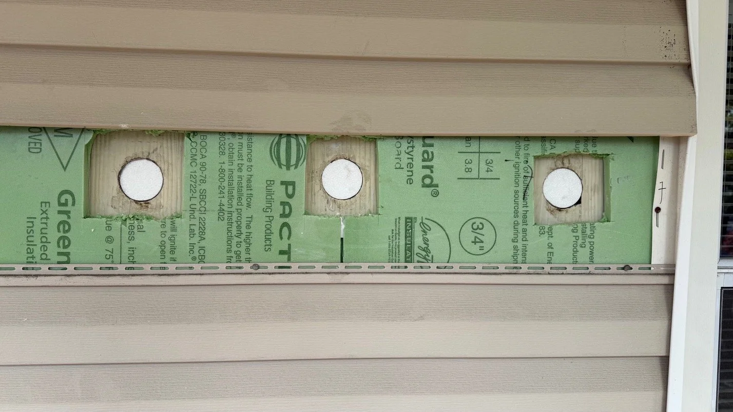 A beige house siding with an open electrical meter box containing green insulation material and three round holes. injection insulation. insulation in existing walls. 