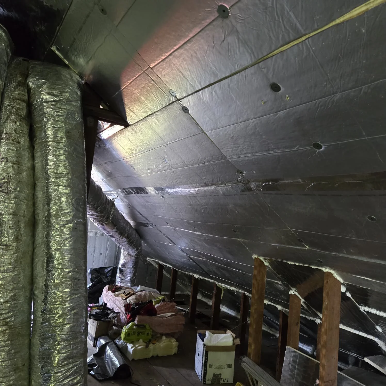 Attic space with unfinished insulation, ventilation ducts, scattered supplies, and exposed wooden beams.