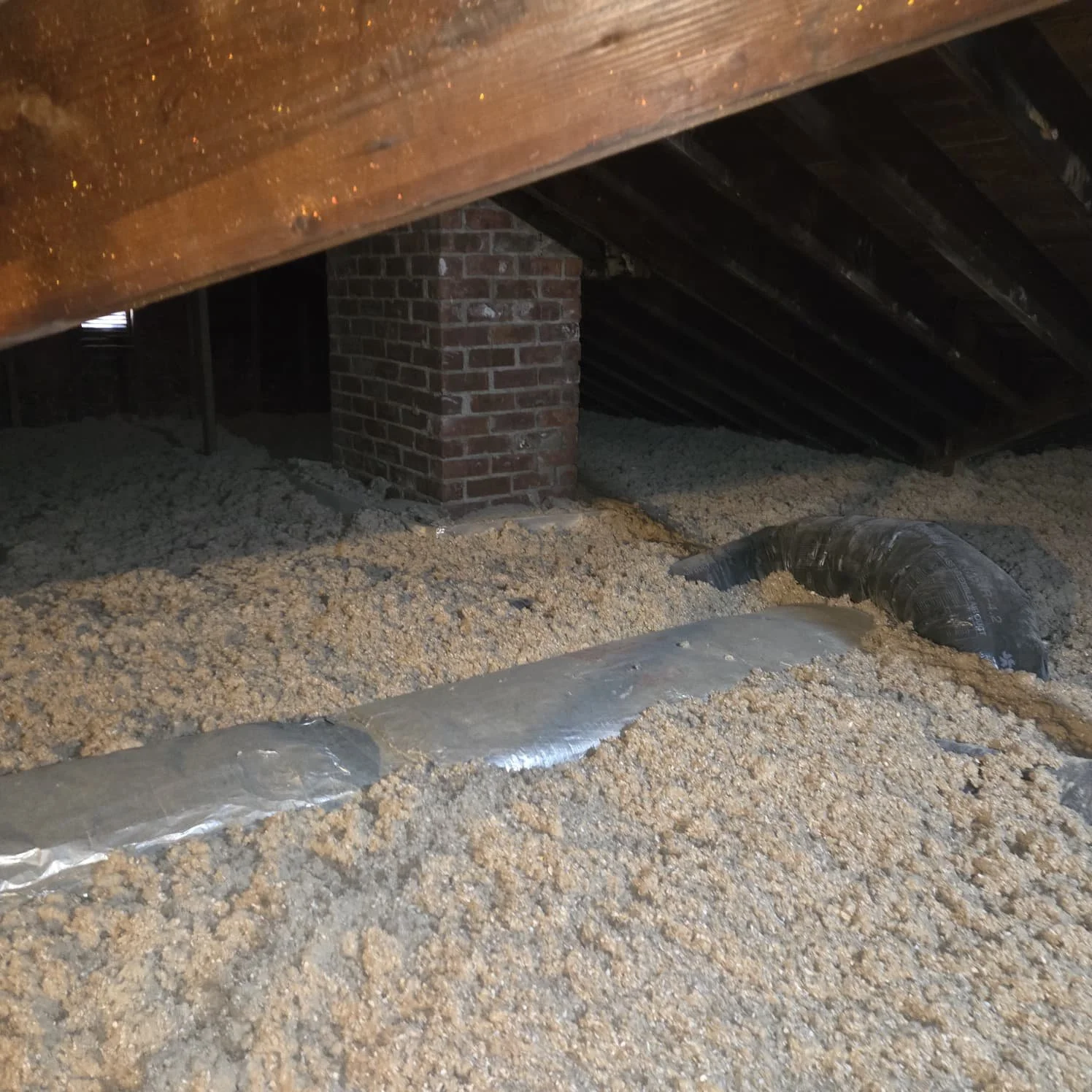 Attic with exposed insulation and ductwork, revealing a brick chimney and wooden roof supports.