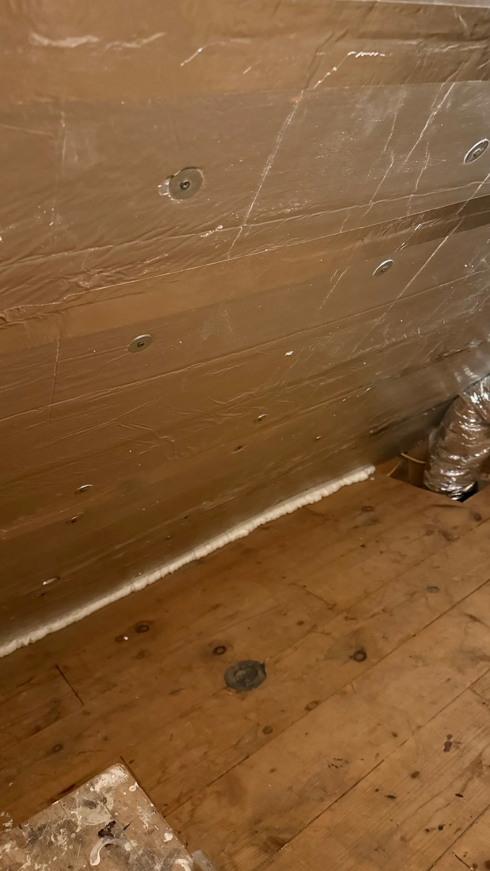 Close-up view of a wooden floor and wall with a white foam insulation line along the base of the wall, and plastic-wrapped duct in the corner.