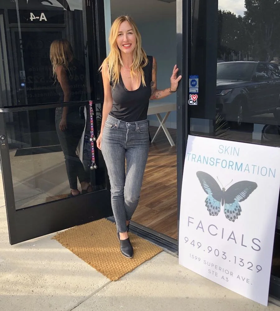 A woman with blonde hair, wearing a black sleeveless top, blue jeans, and black shoes, standing in front of a storefront with a glass door. She is smiling and holding the door open. There is a white signboard next to her advertising skin transformation facials with a butterfly graphic, and a contact phone number. The entrance has a brown doormat.