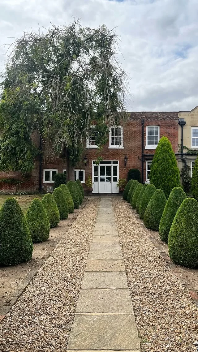 A pathway lined with neatly trimmed green bushes on both sides, leading to a brick house with white-framed windows and a door, under a partly cloudy sky.