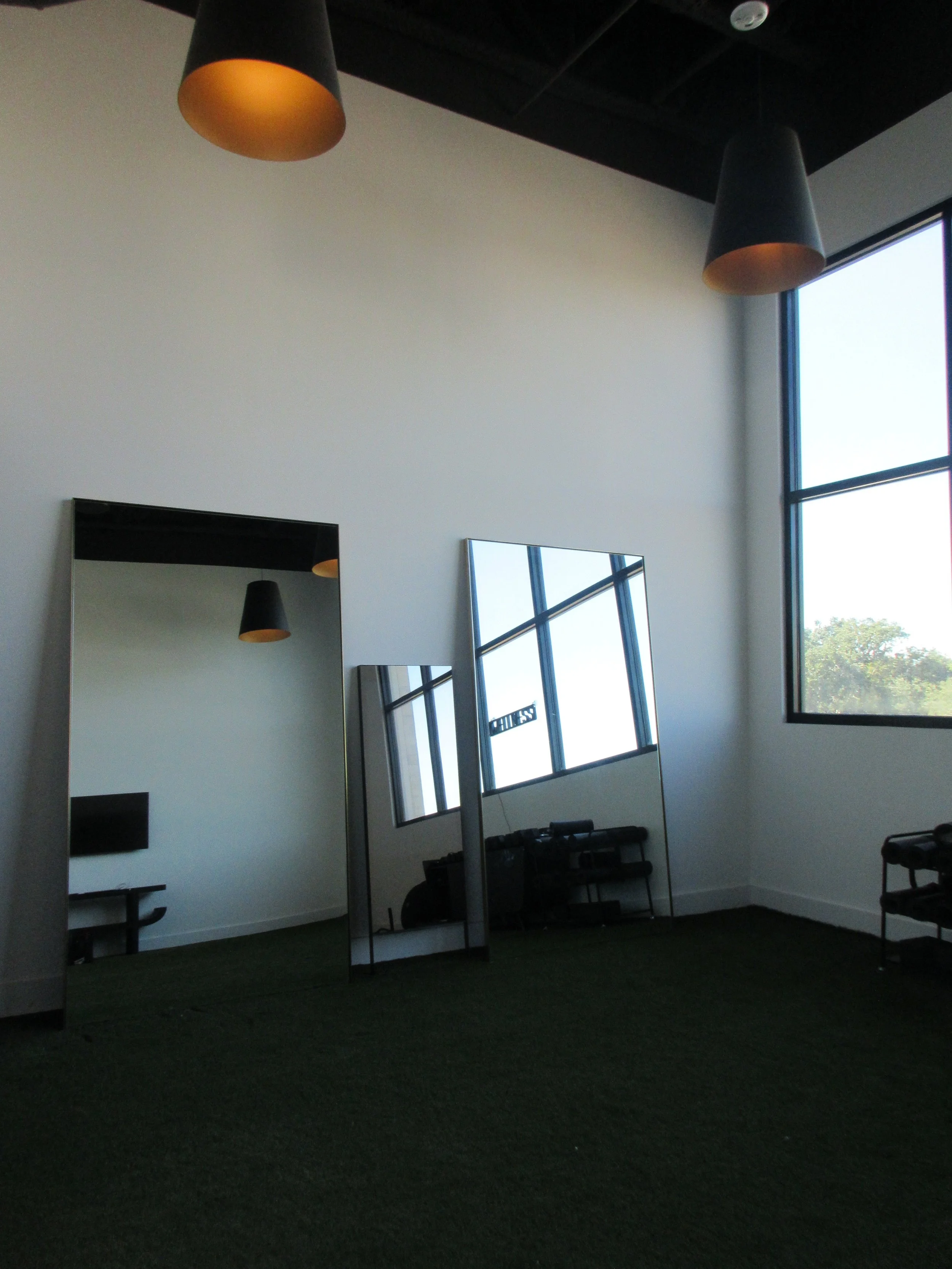 Interior of a room with two black pendant lights hanging from the ceiling, a large window with a view outside, and three mirrors leaning against the wall reflected in the window.