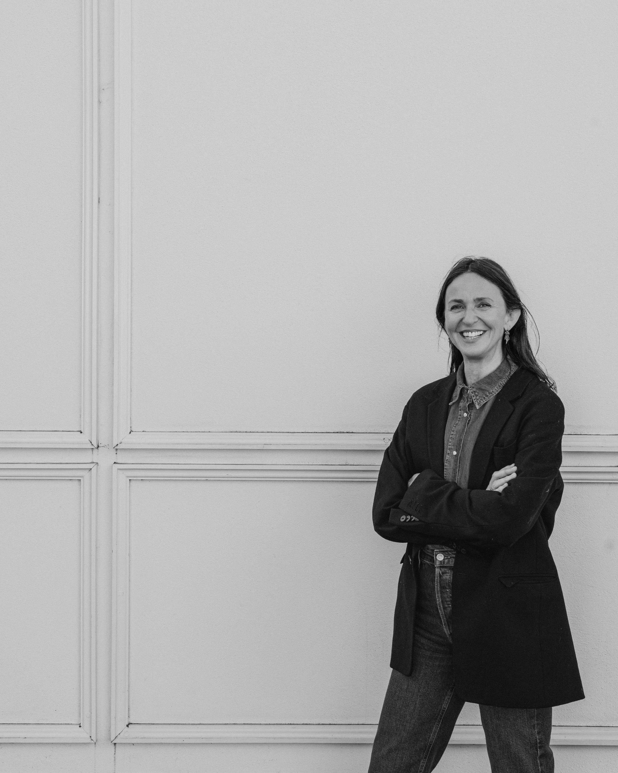Smiling woman in denim shirt and black blazer standing against a cream-colored wall with arms crossed.