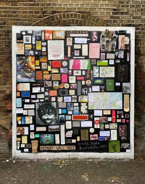 A large outdoor bulletin board filled with various pictures, notes, and artwork, painted on a white background and mounted on a brick wall.